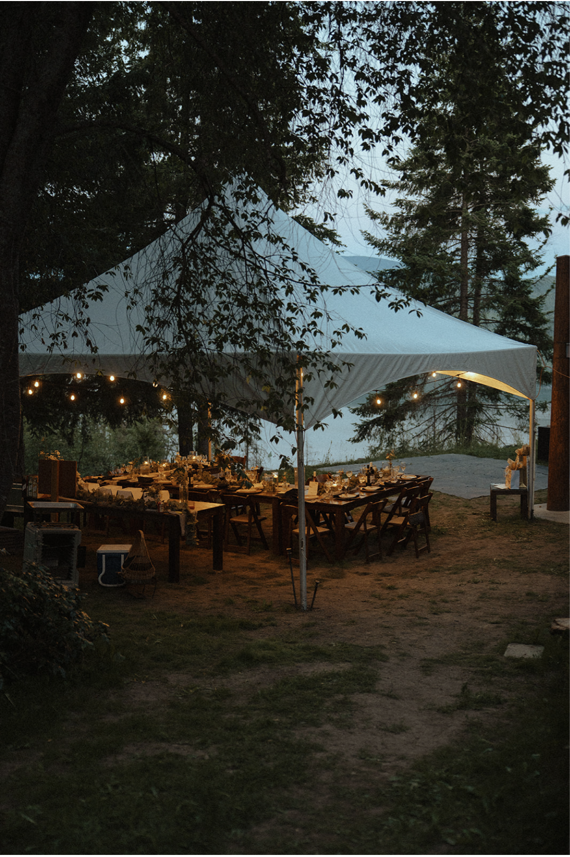 Dinner table set outdoors under a large canopy tent, decorated with string lights, in a forested area at dusk.