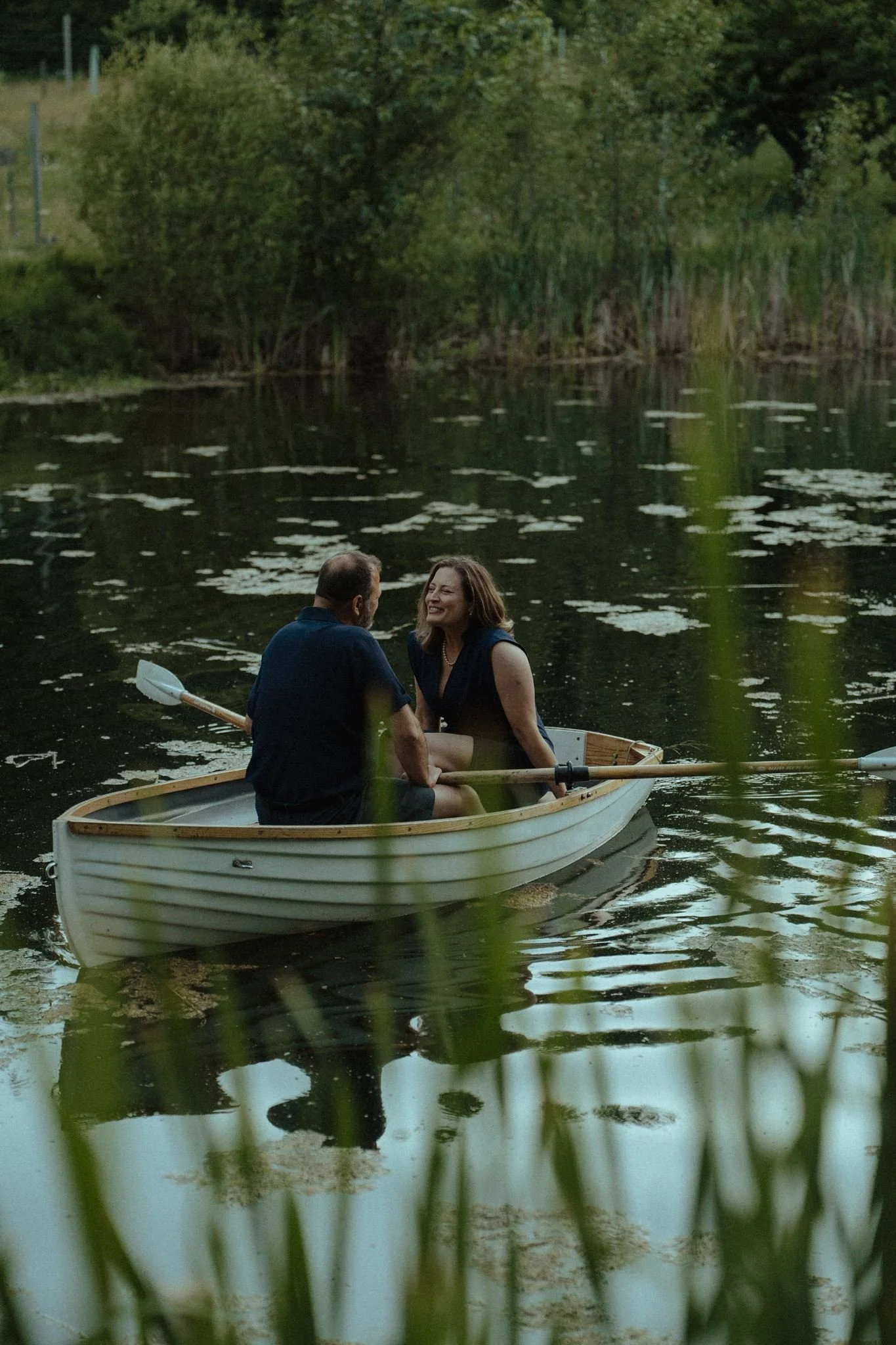 A man and woman in a small boat on a calm lake, surrounded by green trees and reeds, with the woman smiling brightly.
