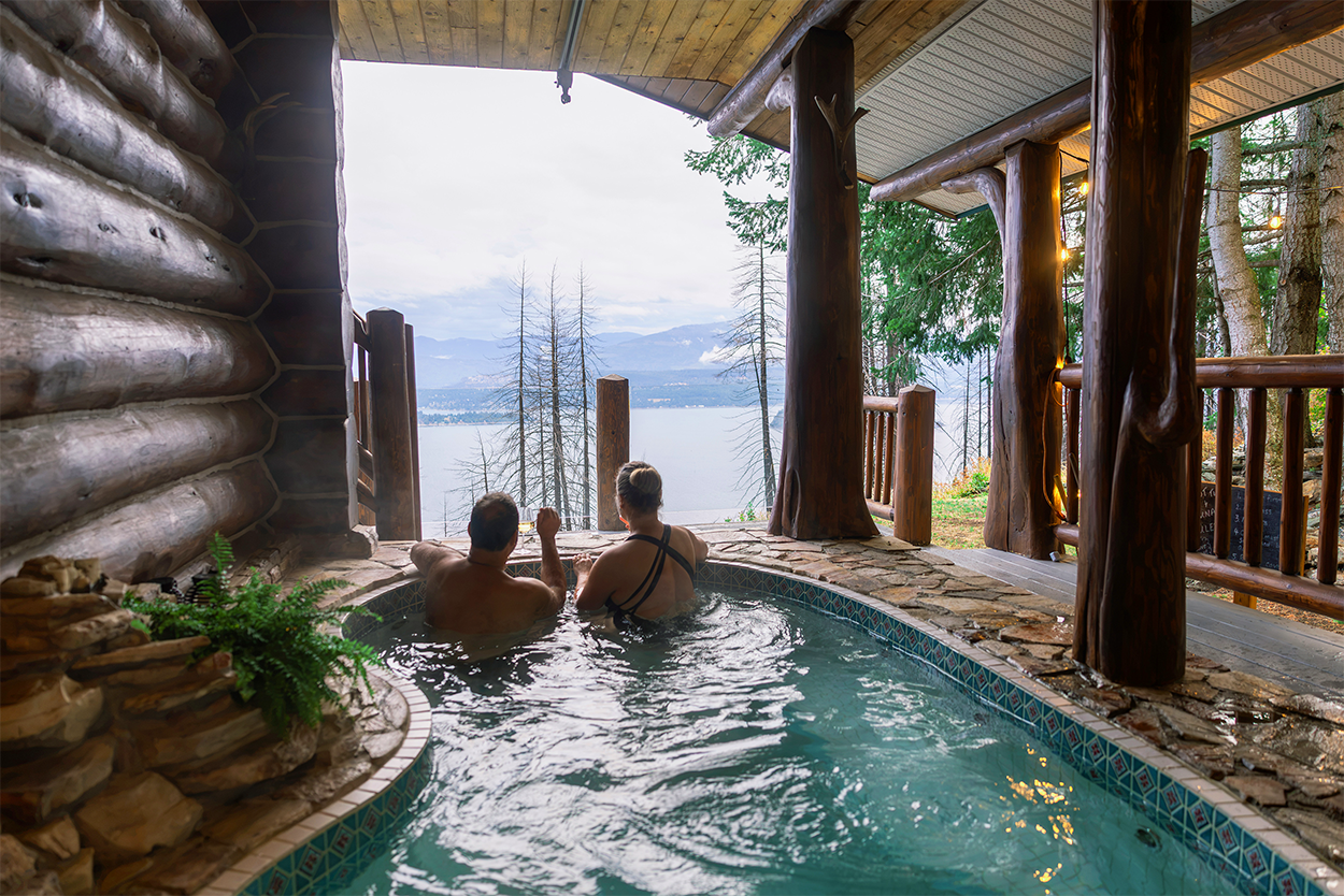 Couple relaxing in outdoor hot tub on a porch, with scenic view of a lake and mountains in the distance, surrounded by trees.
