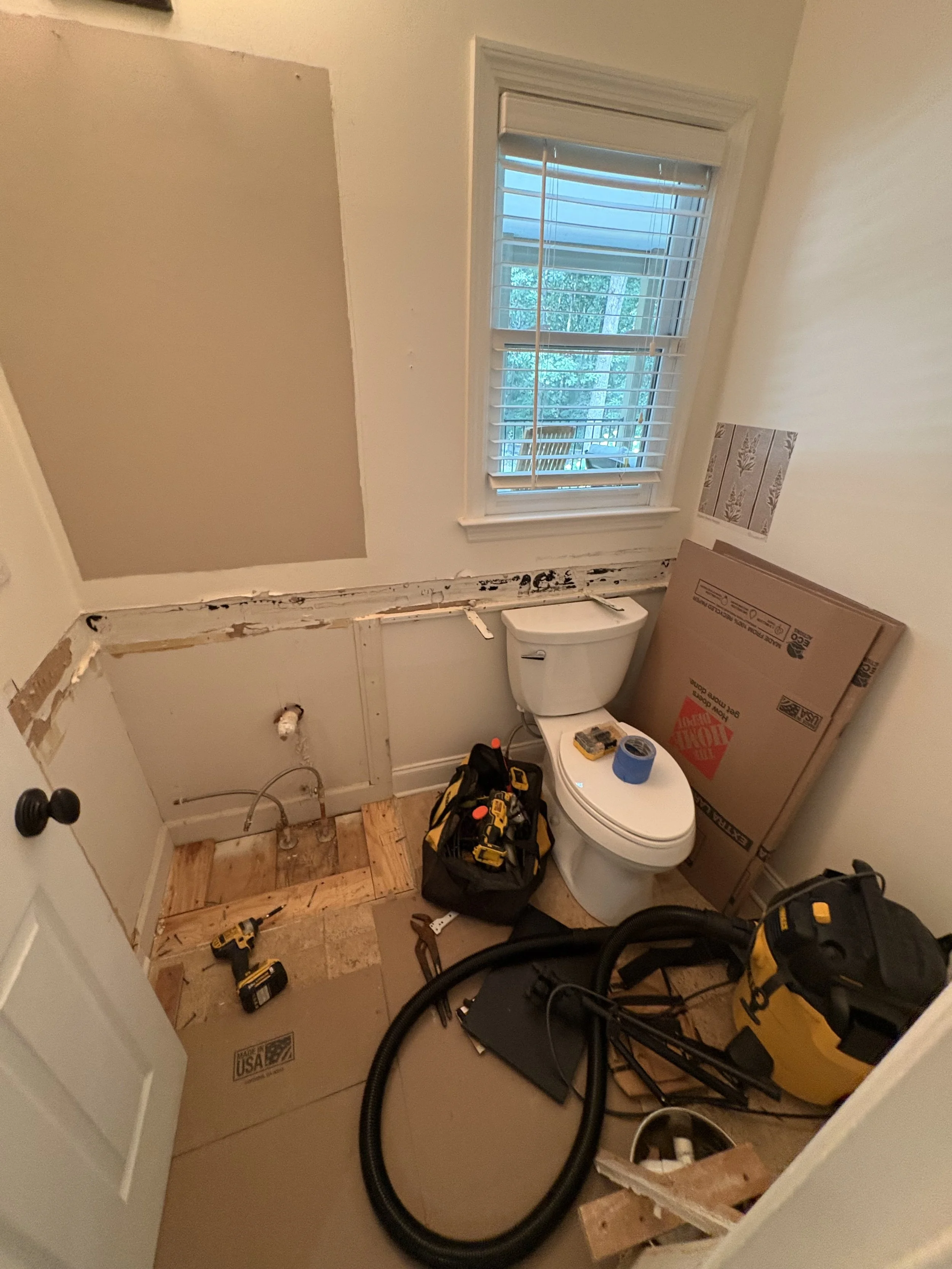 A bathroom under renovation with drywall partially removed, exposing plumbing and wall space. There is a toilet, construction tools, cardboard boxes, and a window with white blinds. The floor has wooden subflooring and construction debris is visible.