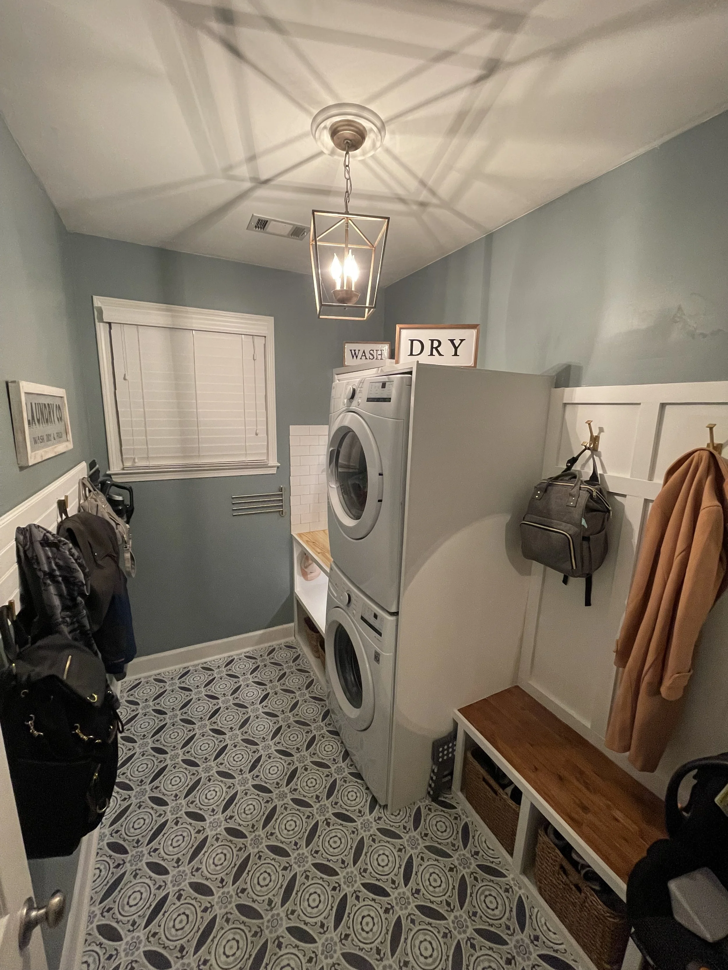 A laundry room with a patterned tile floor, white shelves with baskets, and a stacked washer and dryer. Walls are painted light gray, with coats hanging on hooks. There are signs above the appliances that say "Wash" and "Dry."