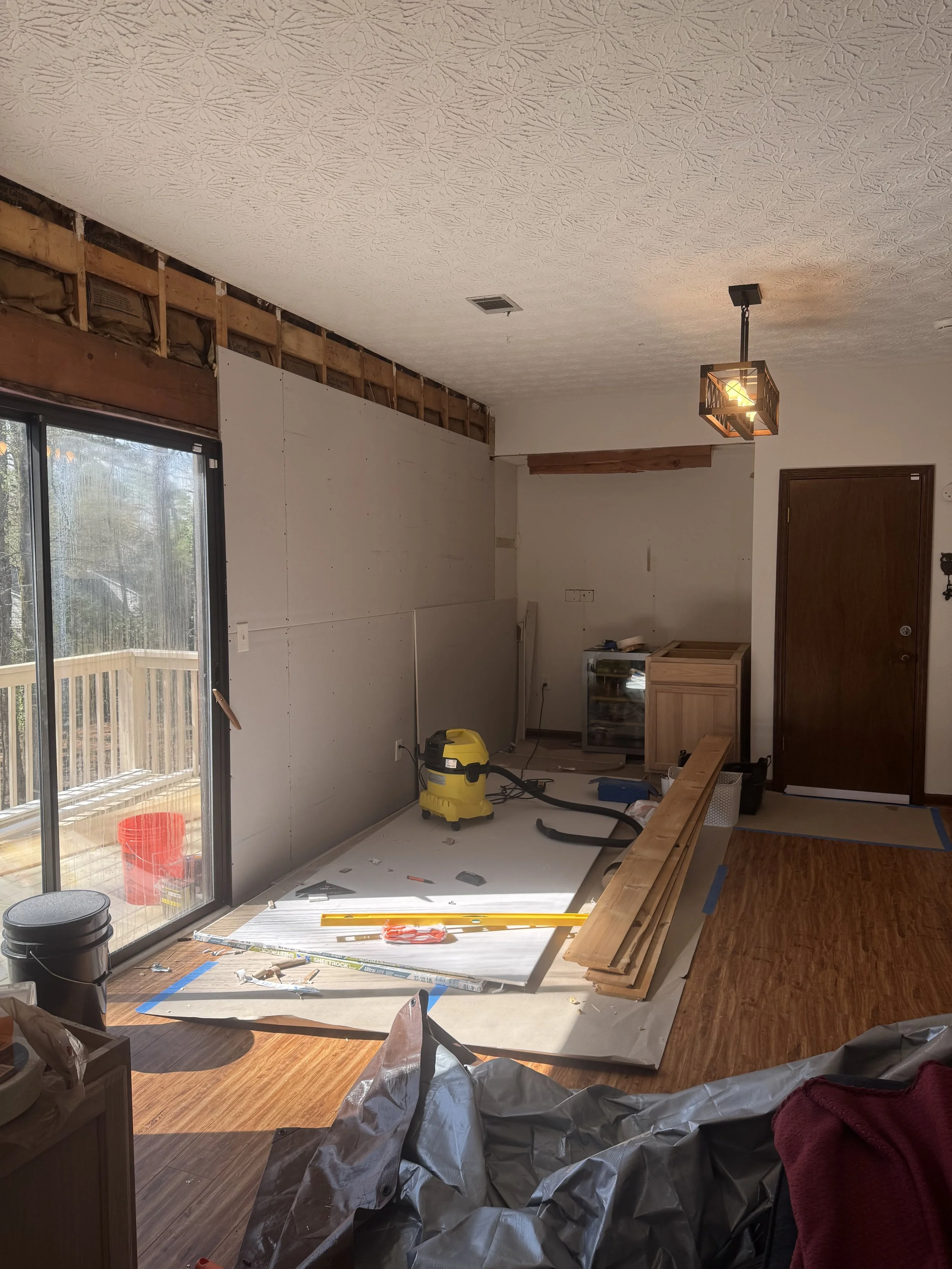 Living room under renovation with exposed wall framing, construction tools, wooden flooring, and partially completed wall panels.