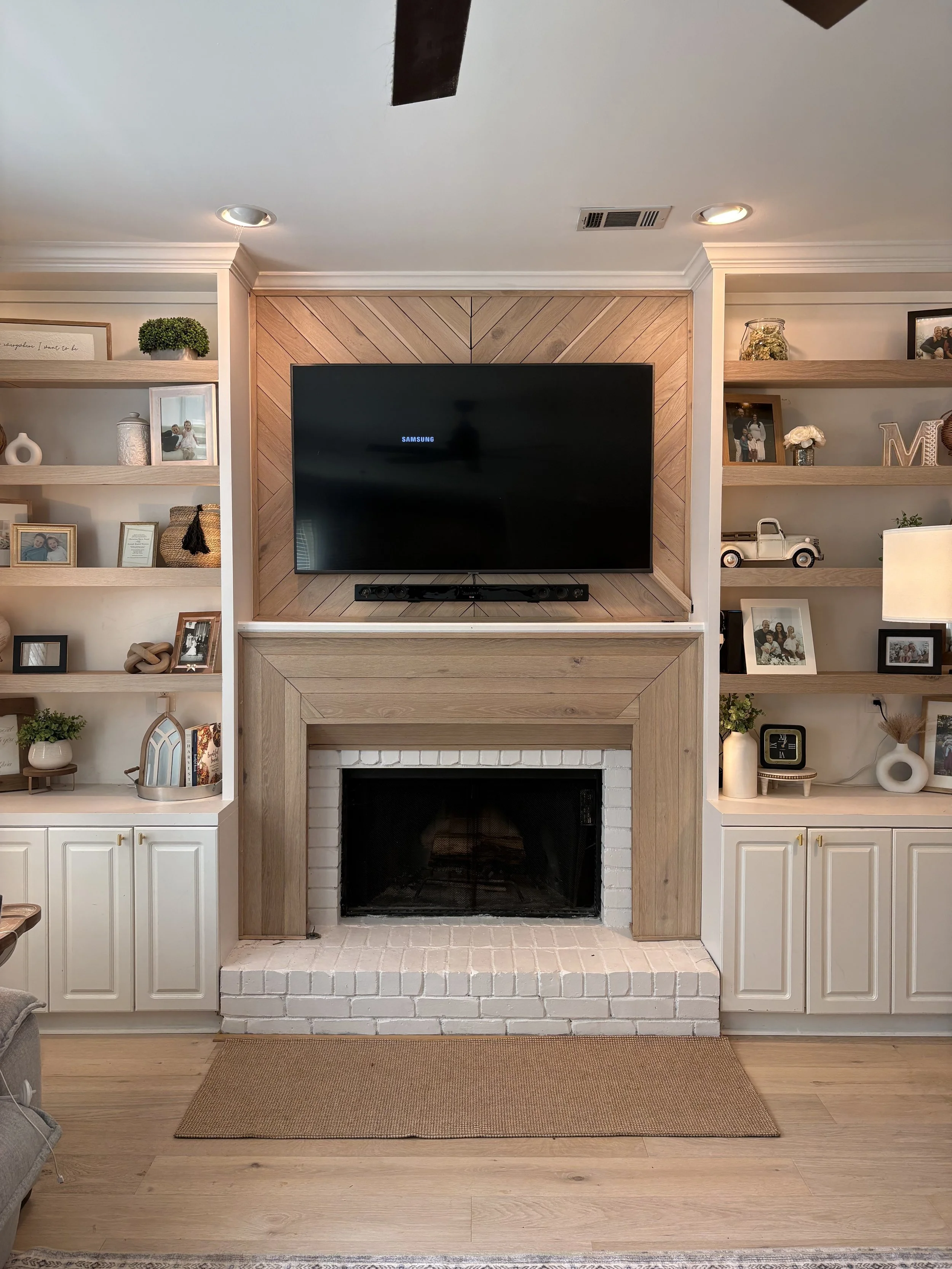 Living room fireplace with mounted TV above, white brick surround, wooden mantel, and built-in shelves filled with framed photos, decorative items, books, and plants. Hardwood floor with a beige rug.