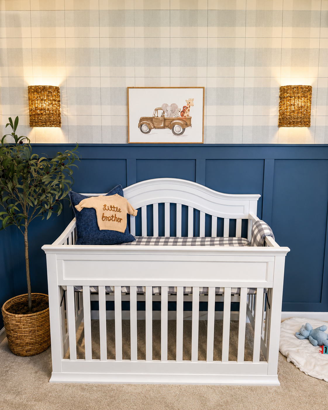 A white baby crib with a checkered mattress in a nursery with blue and cream walls. A navy pillow with a cream knit sweater labeled 'little brother' rests on the crib. To the left, there's a potted plant, and above, a framed picture of animals in a truck. Two woven wall lights are mounted above the crib.