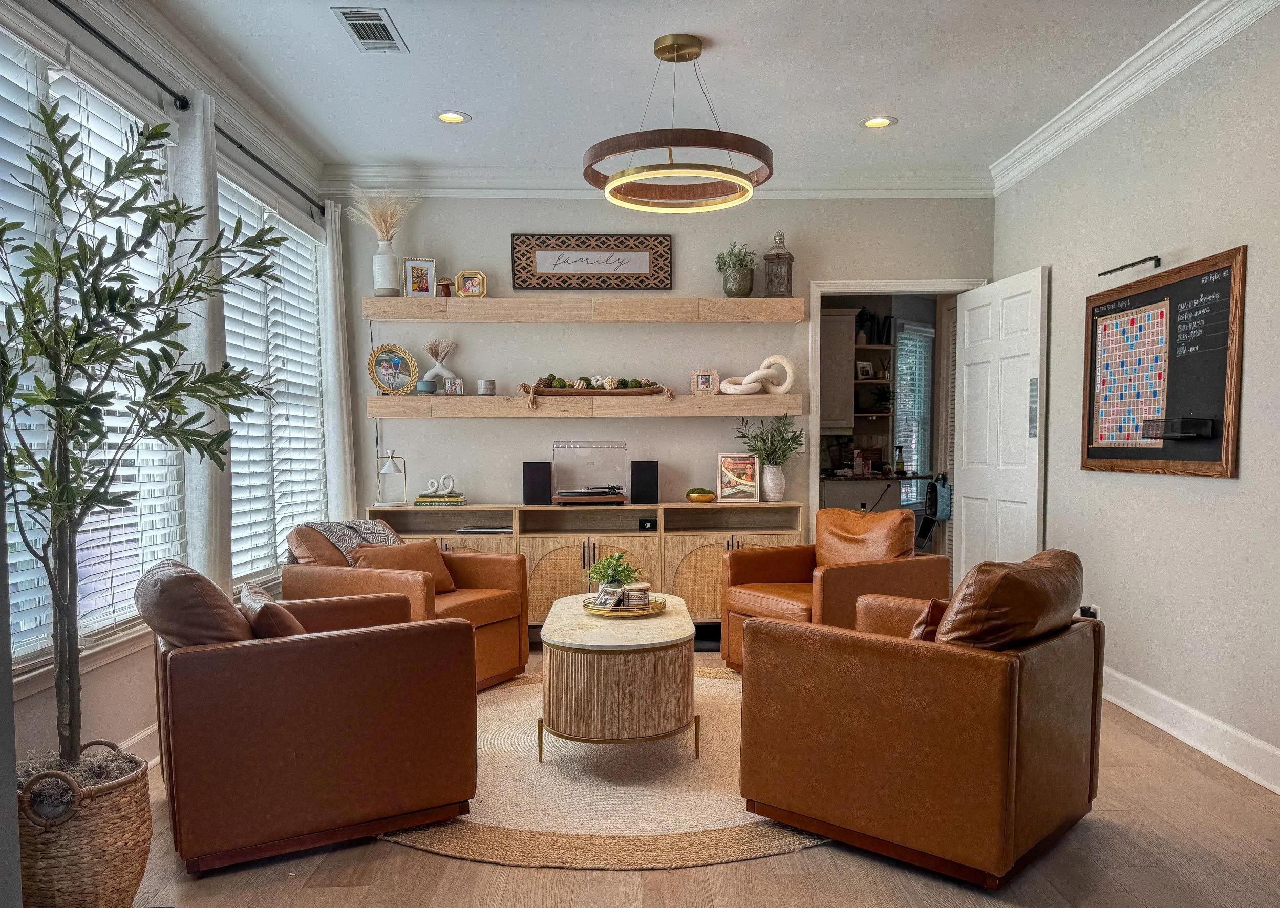 A living room with a round wooden table, several leather armchairs, a large window with blinds, and two floating wooden shelves decorated with plants, pictures, and decorative items. A modern circular chandelier hangs from the ceiling.
