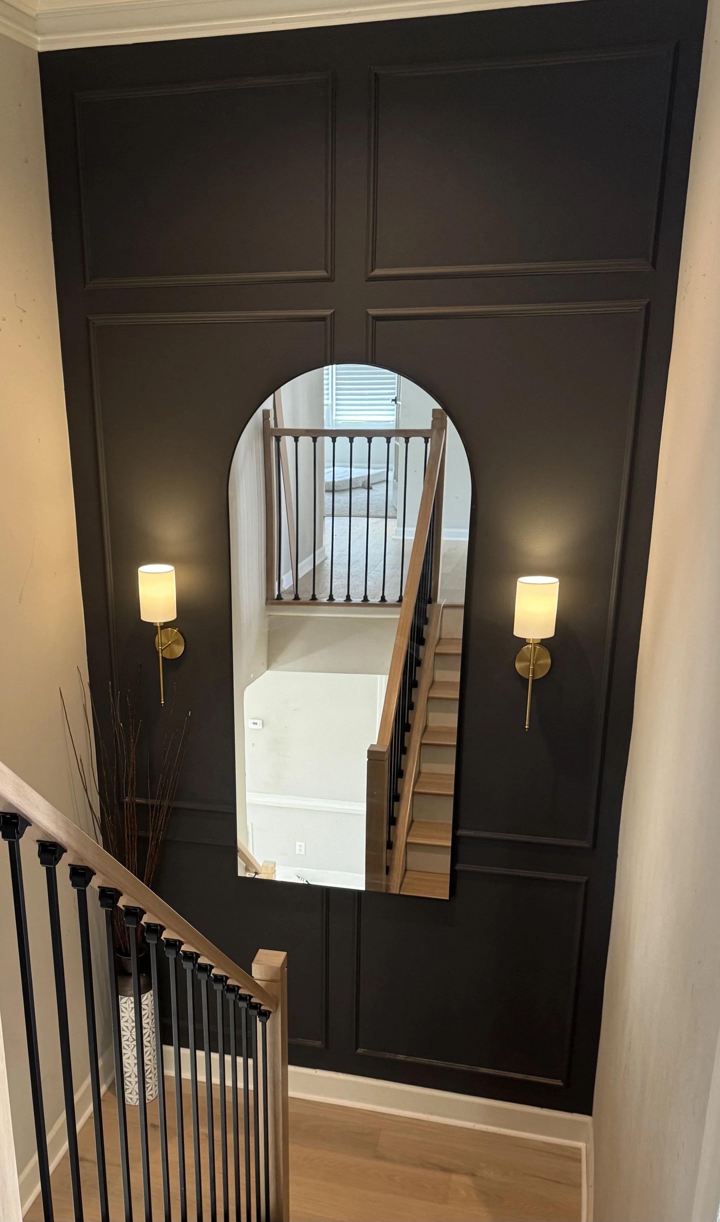 Interior view of a house staircase area with dark wall paneling, a large mirror reflecting a staircase and railing, two wall-mounted lights on either side of the mirror, and a decorative vase with tall sticks in the corner.