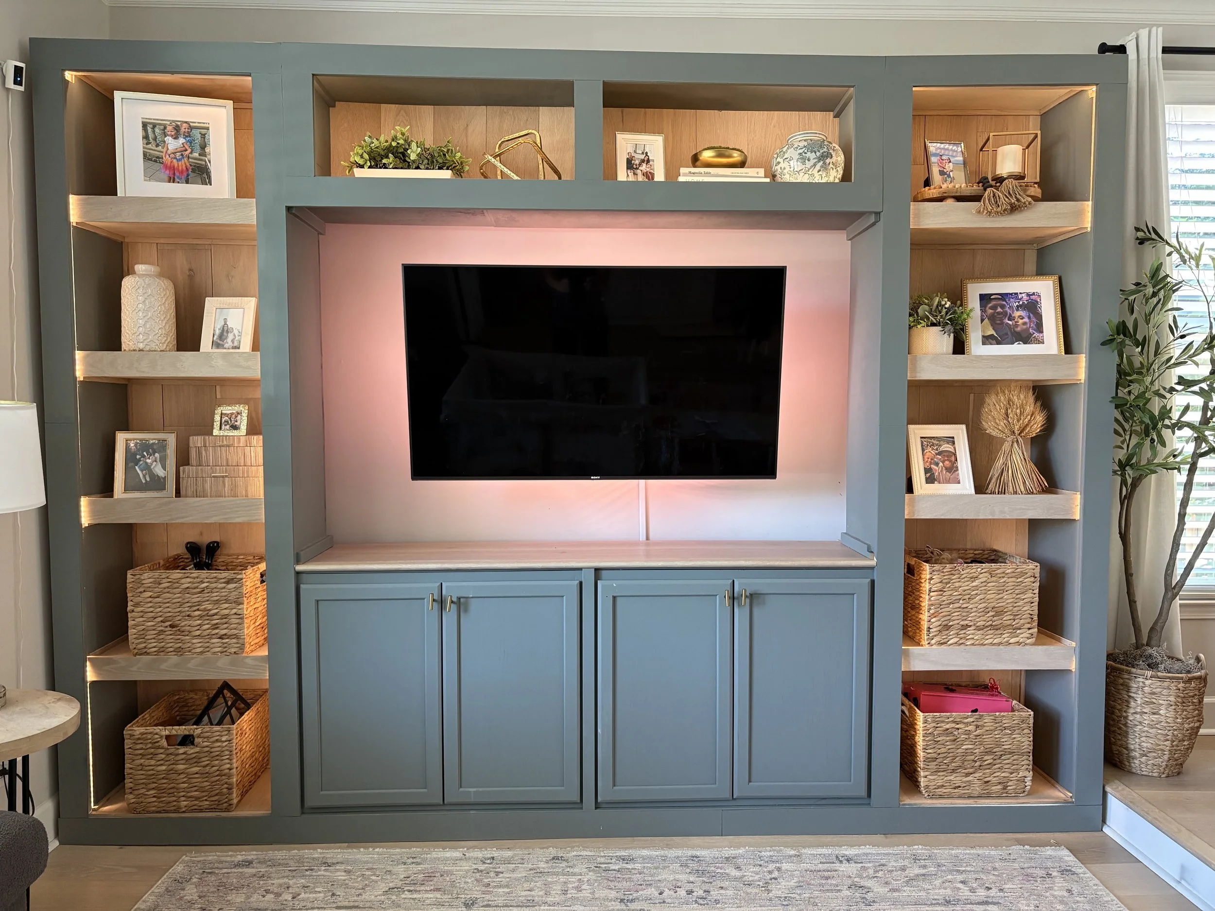 A blue entertainment center with open shelves decorated with framed photographs, books, plants, and decorative objects. The center surrounds a flat-screen television, and woven baskets are placed on the lower shelves.