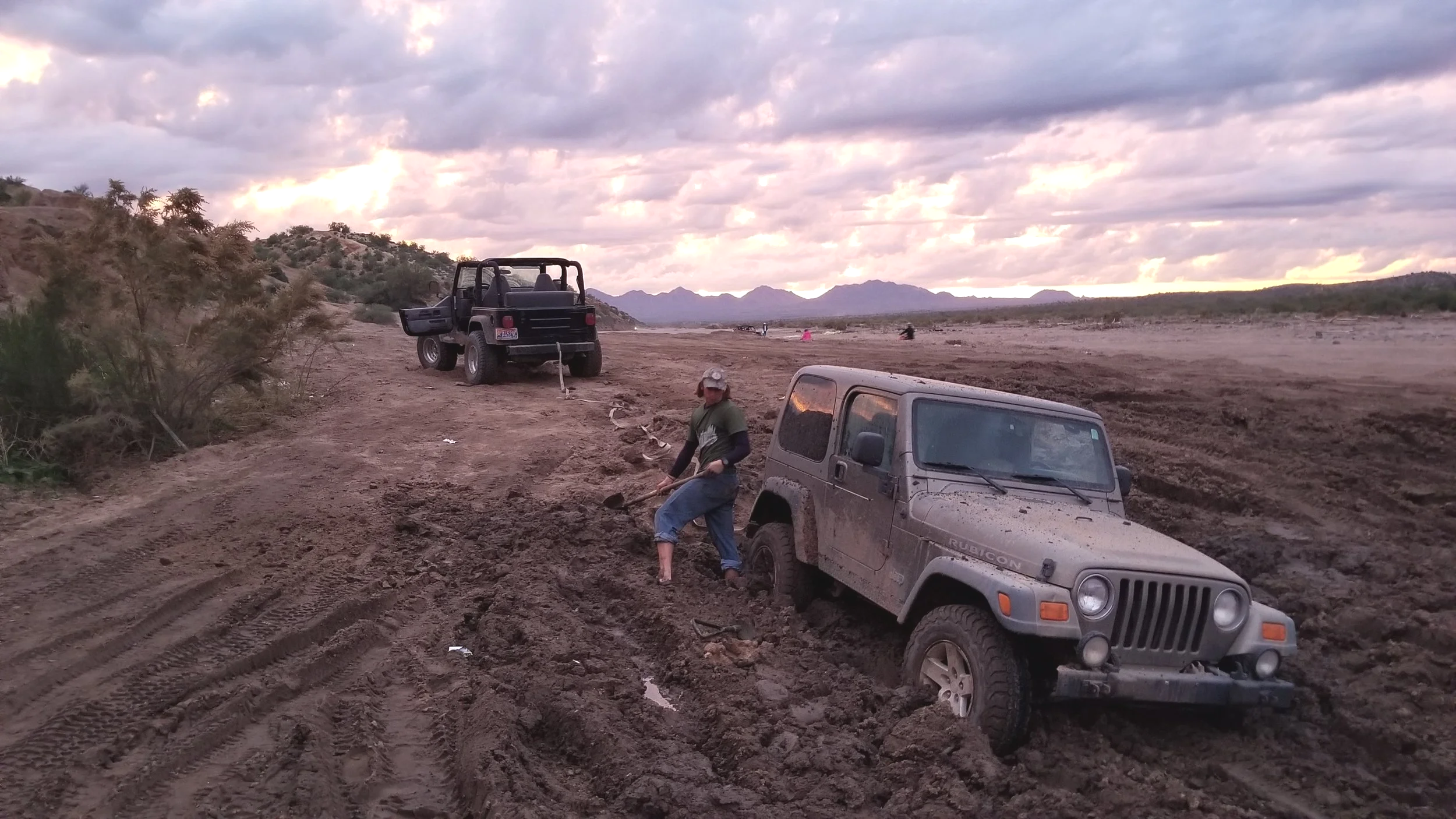 A muddy off-road scene with two vehicles, one parked and one partially stuck, and a person shoveling mud near the front of a Jeep Rubicon with its door open. The landscape features a dirt trail, sparse desert vegetation, and mountains in the distance under a cloudy sky at sunset.