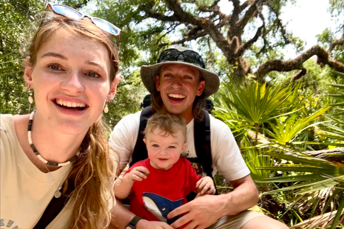 A smiling family of three taking a selfie outdoors in a lush, green jungle setting. The woman is on the left, the man is in the middle holding a young boy, all enjoying a sunny day with trees and plants in the background.