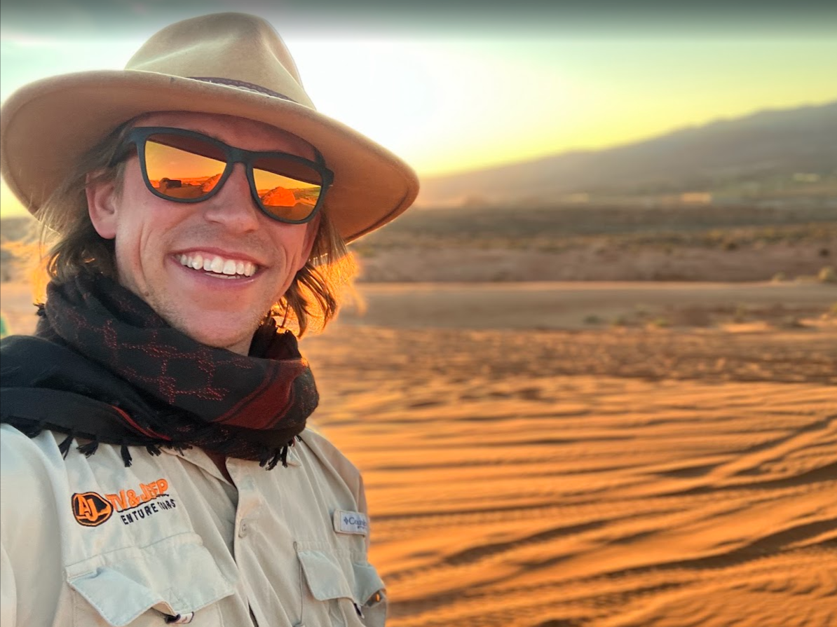 A man outdoors at sunset, smiling and wearing a wide-brimmed hat, sunglasses with orange reflection, a light-colored shirt with logo patches, and a black scarf. The background shows a desert landscape with sand dunes and distant mountains.