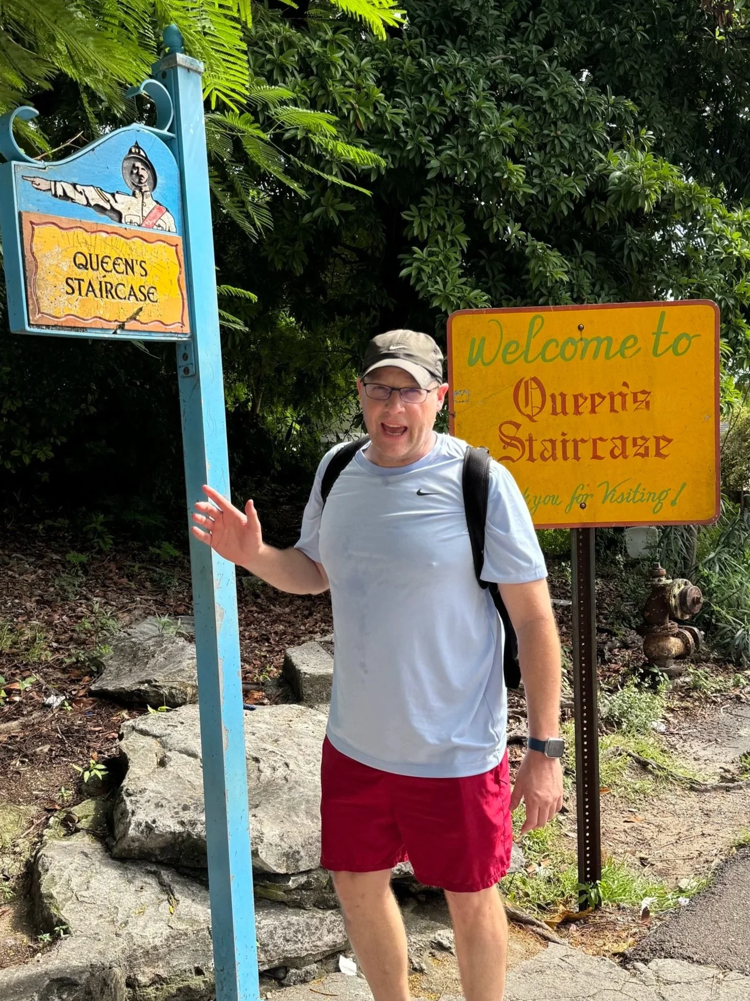 A man wearing a gray cap, glasses, a white shirt, and red shorts is standing outdoors next to a sign that reads "Queen's Staircase" and a yellow sign that says "Welcome to Queen's Staircase" in the Bahamas.