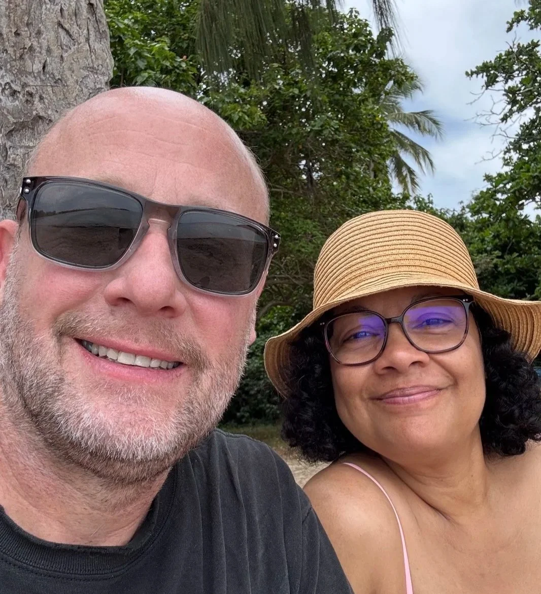 Smiling Gartz Hanson with sunglasses and a smiling wife, Rosa Moreno, with sunglasses and a wide-brimmed hat taking a selfie outdoors, with palm trees and greenery in the background.