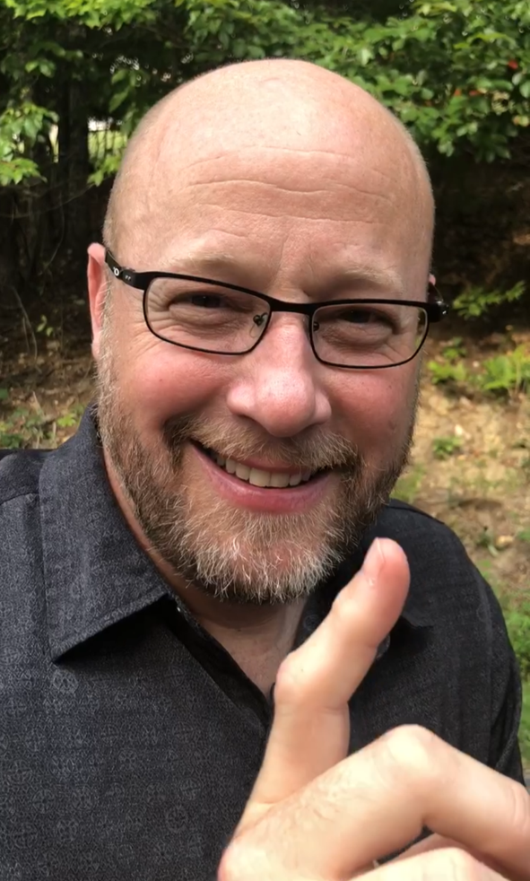 A smiling middle-aged man with glasses and a beard pointing upward outdoors with green leaves in the background.