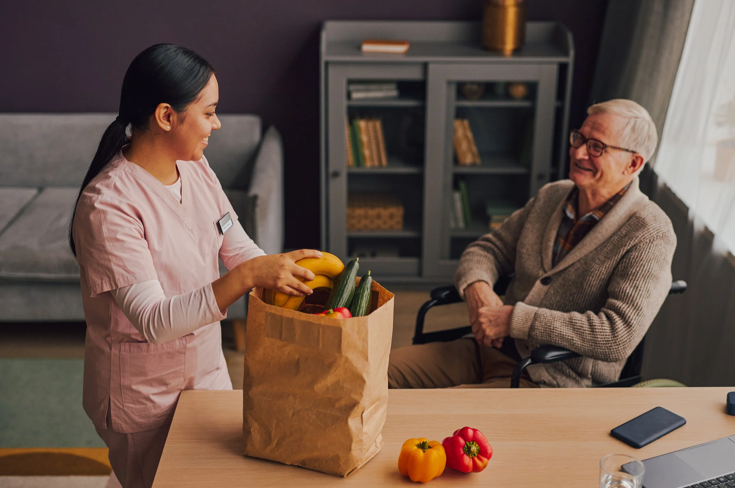 A young woman in pink scrubs hands a paper bag filled with fresh vegetables to an elderly man sitting at a table, with a few bell peppers placed on the table.