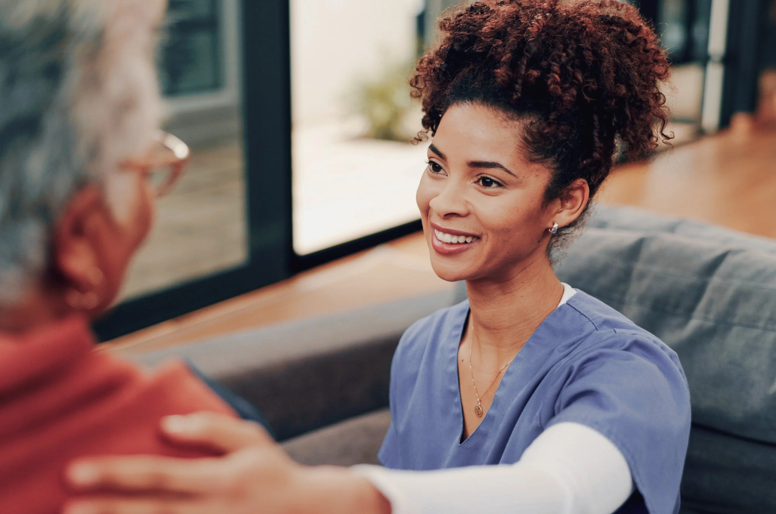 A young woman in medical scrubs smiling and talking with an elderly person, setting appears to be in a healthcare or caregiving environment.