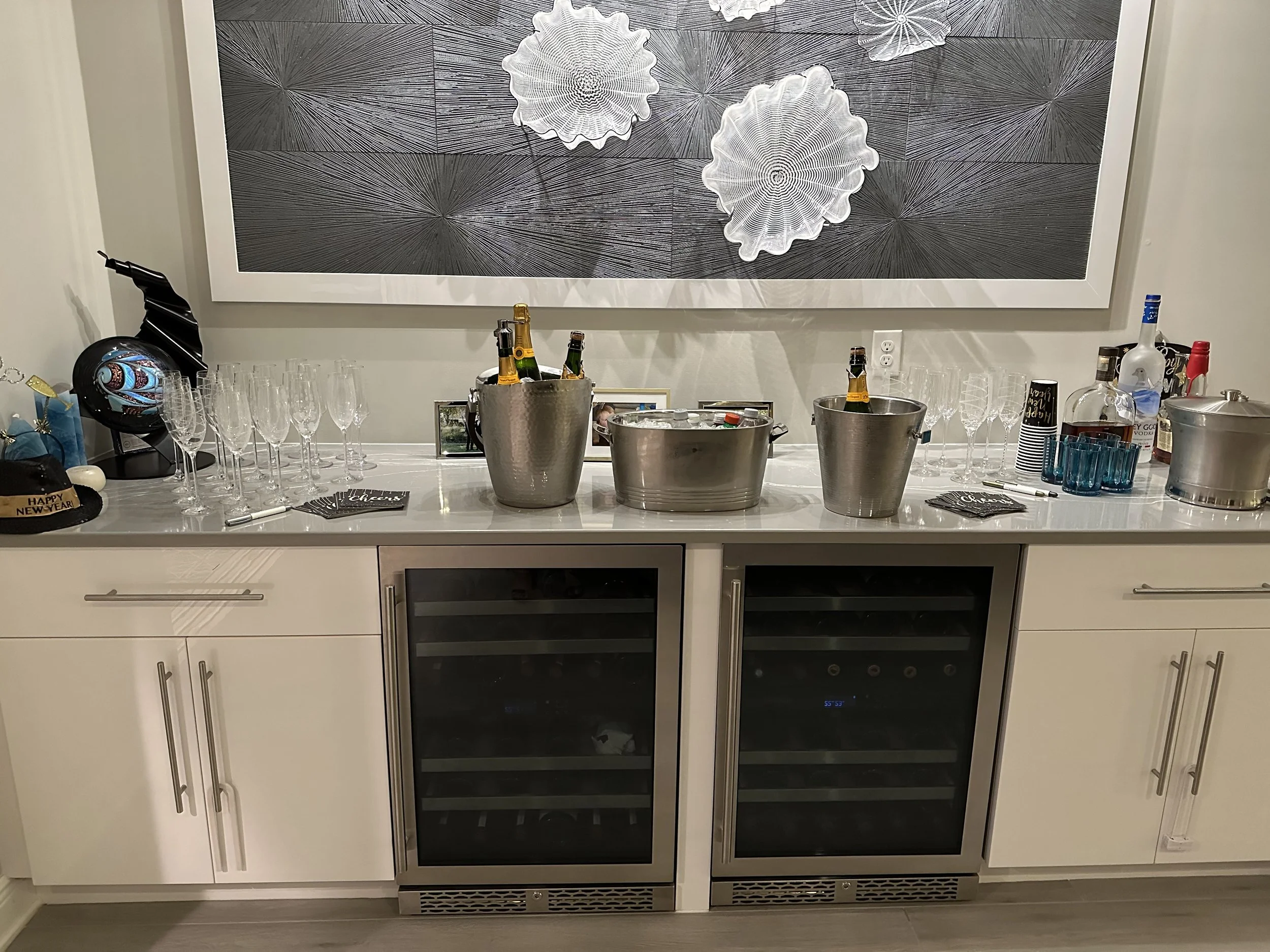 A kitchen countertop with empty champagne glasses, beer bottles in ice buckets, a bottle of vodka, and bar accessories, with abstract wall art behind.