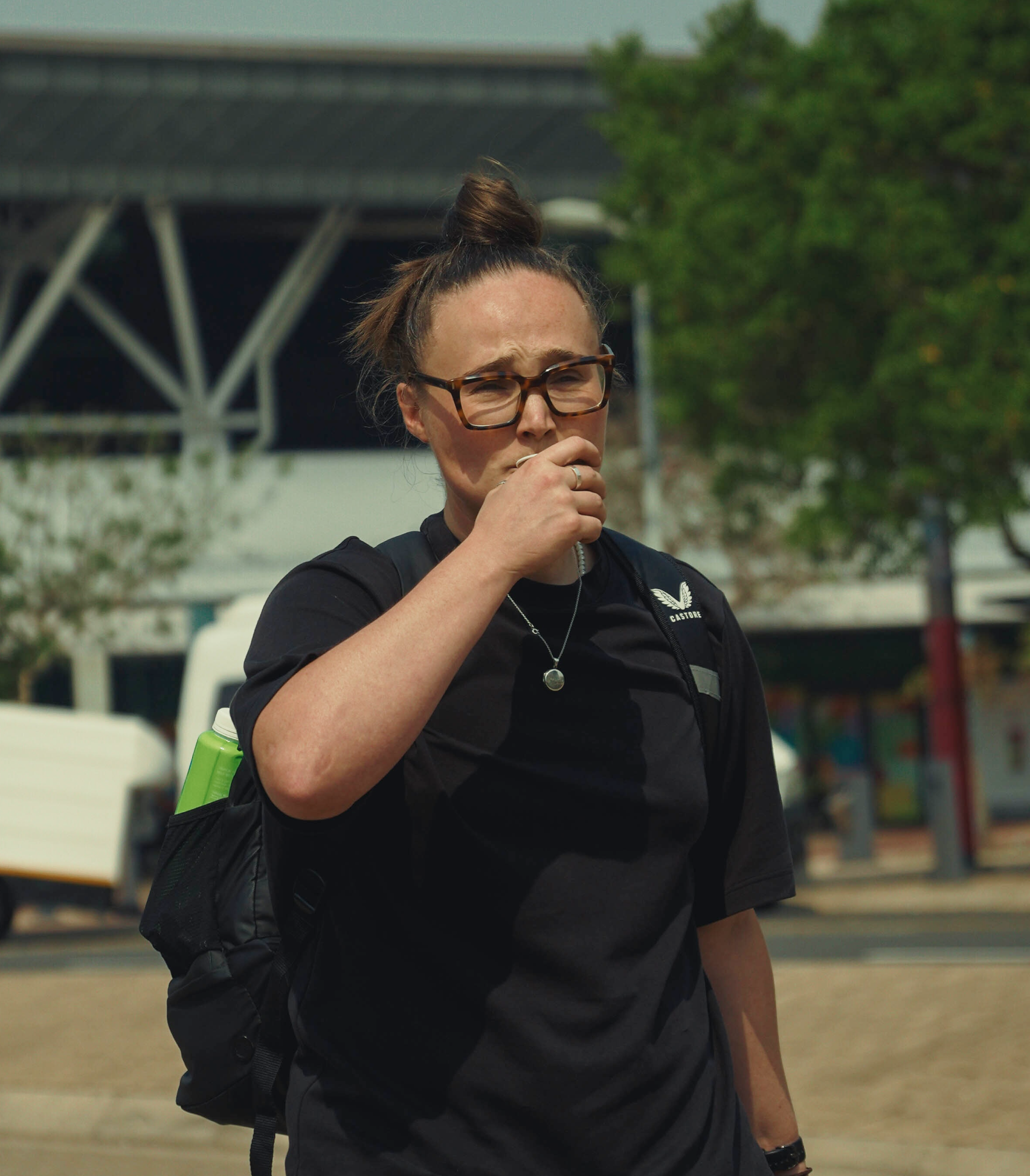 A woman with glasses and a bun hairstyle, wearing a black uniform with a badge, holding her hand near her mouth in an outdoor setting with trees and a bridge in the background.