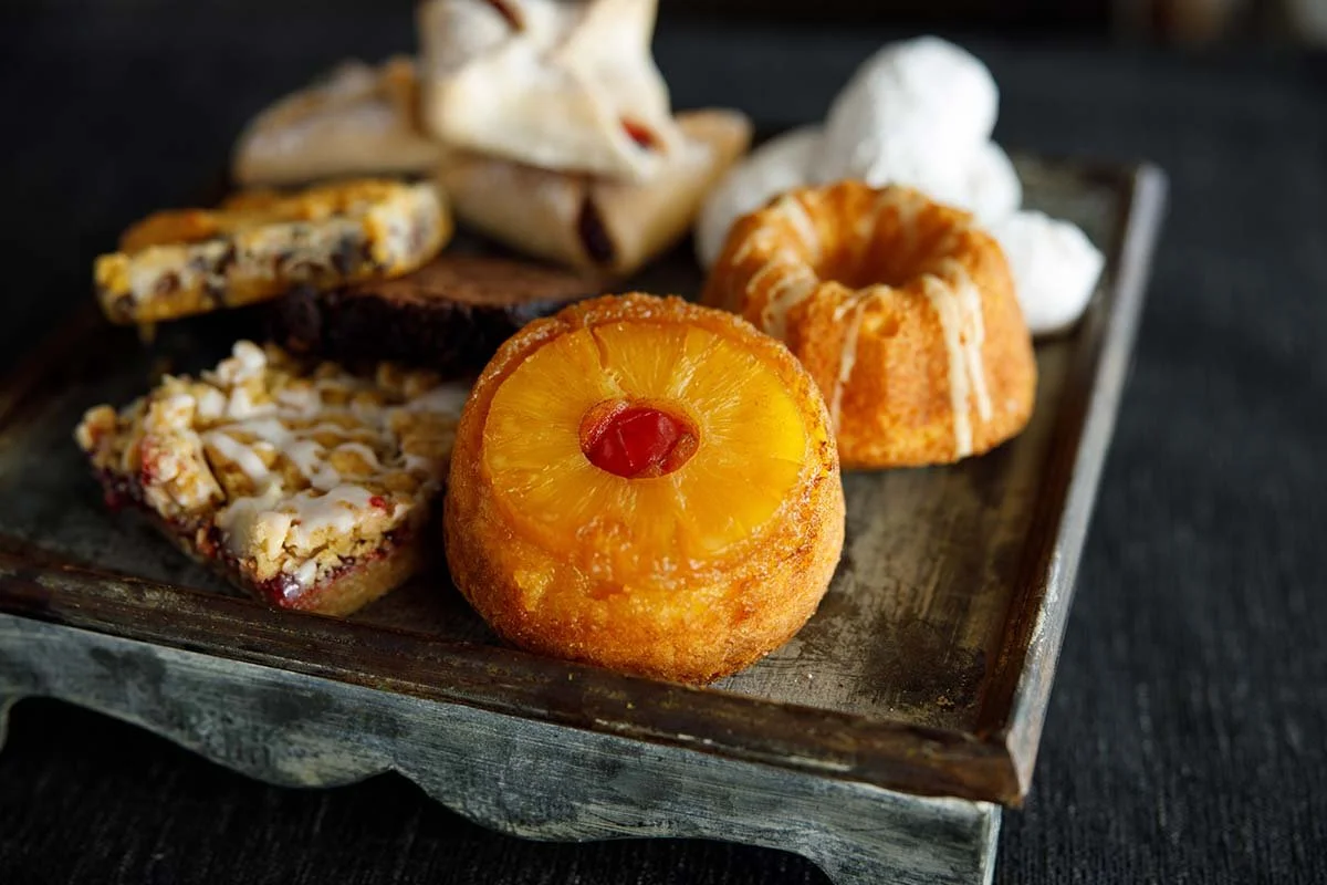 Assorted pastries and cookies on a rustic serving tray, including pineapple upside-down cake with a cherry and a glazed Bundt cake.