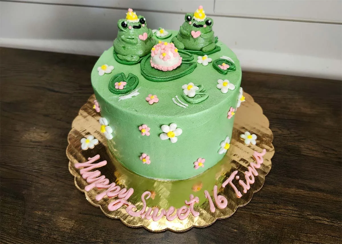 Green frog-themed birthday cake decorated with flowers, lily pads, and three party-hat frogs
