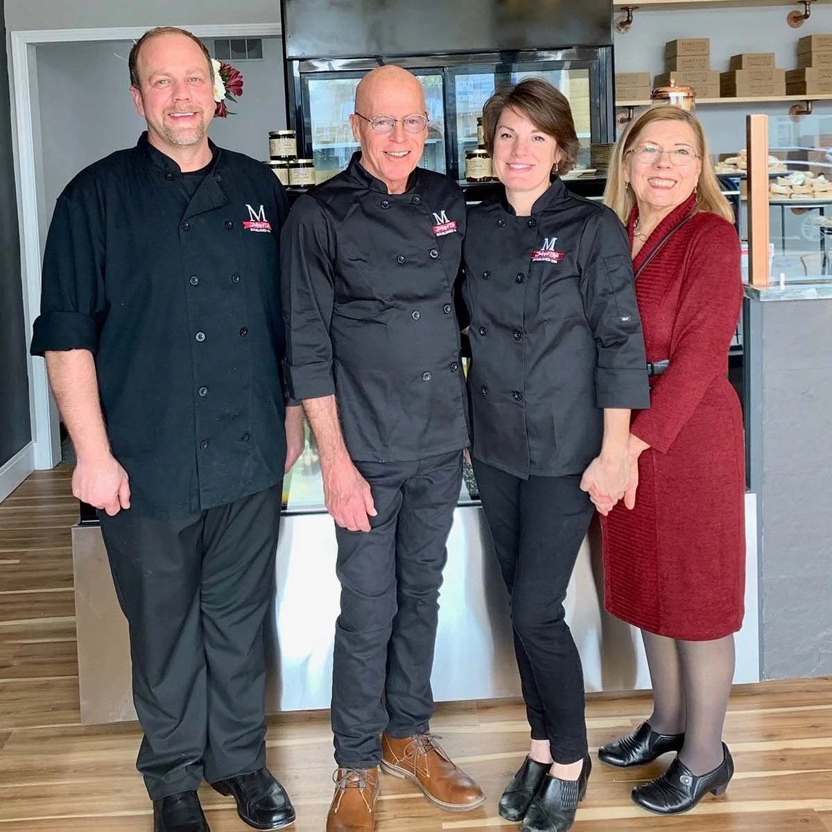 Four people, including three chefs in black uniforms and a woman in a red dress, pose inside a bakery or confectionery shop.