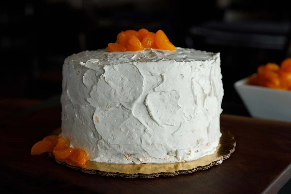 Round white-frosted cake topped and garnished with orange segments on a gold cake board