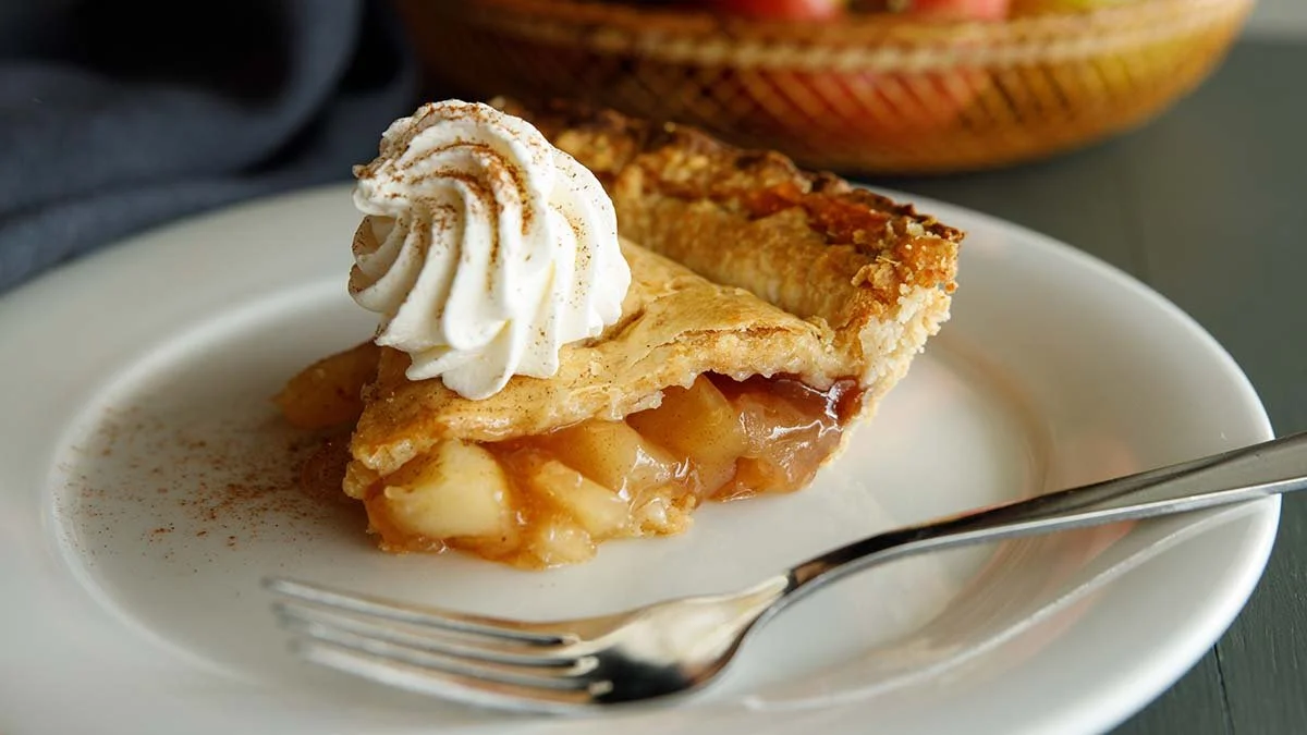 Slice of homemade apple pie topped with whipped cream and cinnamon on a white plate with a fork