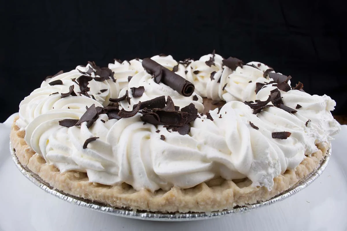 Cream pie topped with whipped cream rosettes and dark chocolate shavings in a foil pie tin