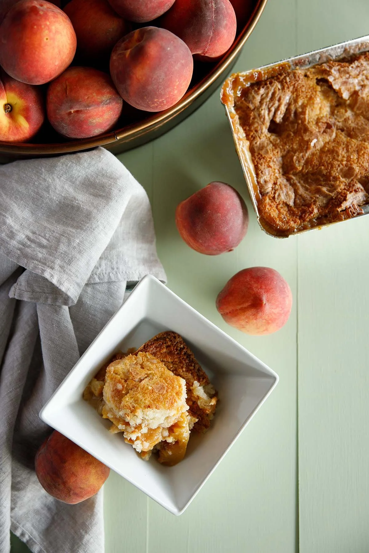 Peaches, peach crisp in a white bowl, and a baking dish of peach dessert on a green table.