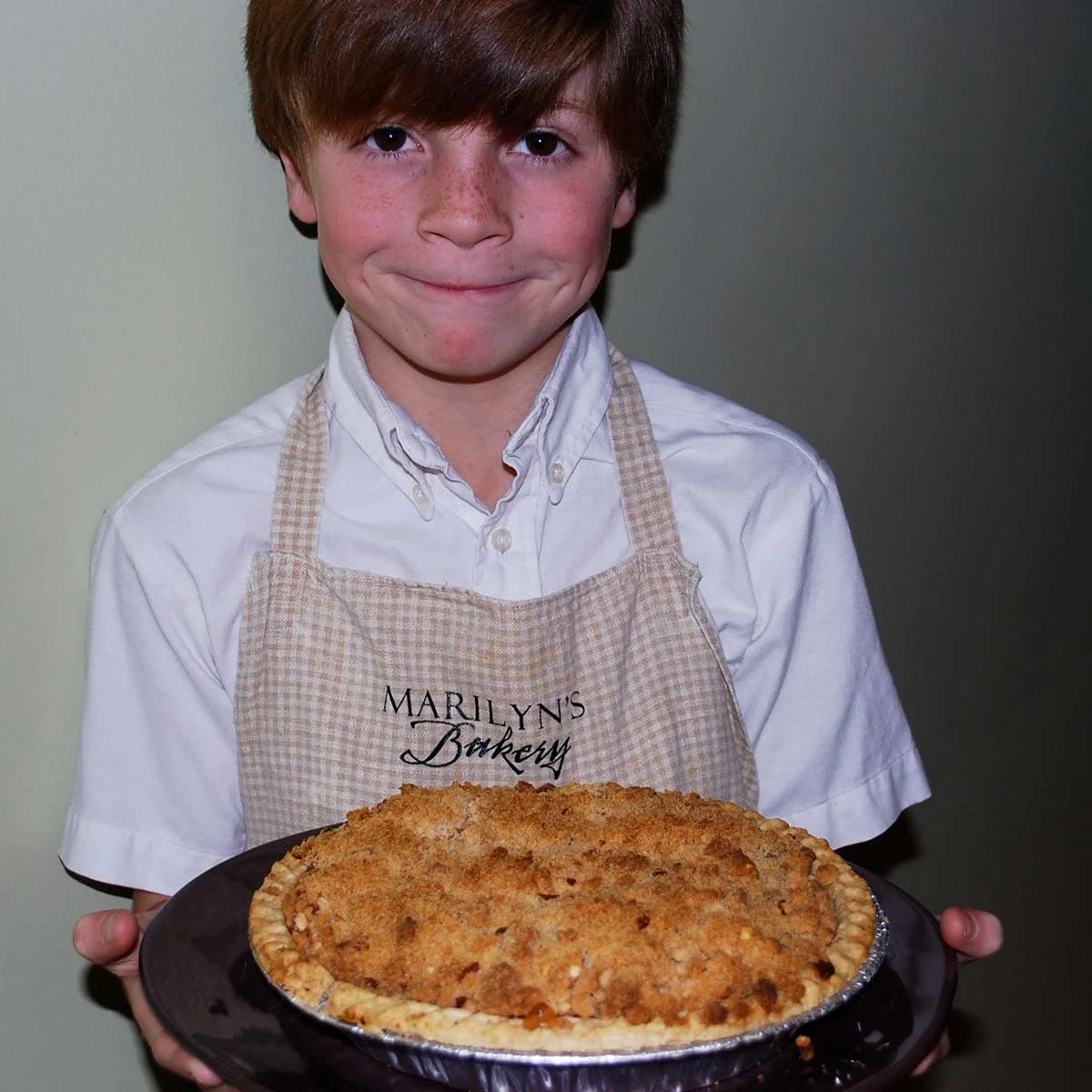Child wearing a Marilyn's Bakery apron holds a large freshly baked crumb-topped pie.