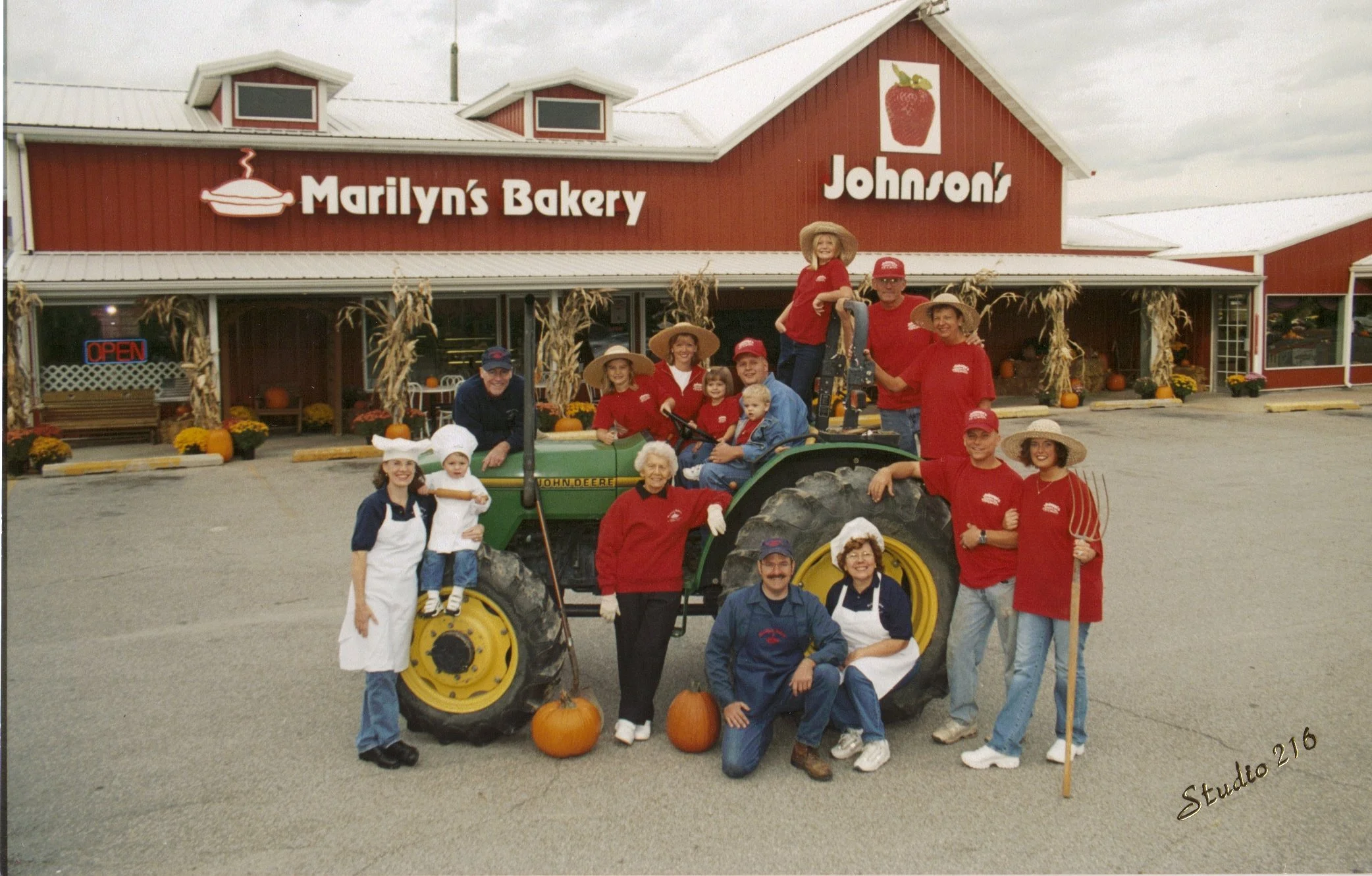 Family group posing with a green John Deere tractor outside Marilyn's Bakery and Johnson's farm market, decorated with pumpkins and cornstalks.