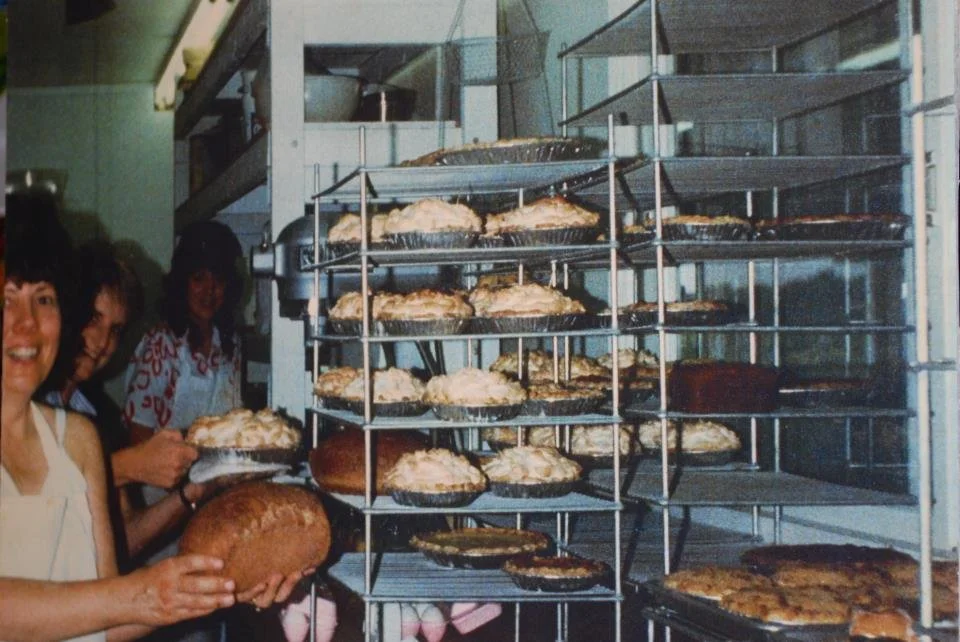 People holding freshly baked pies beside racks filled with homemade pies and baked goods