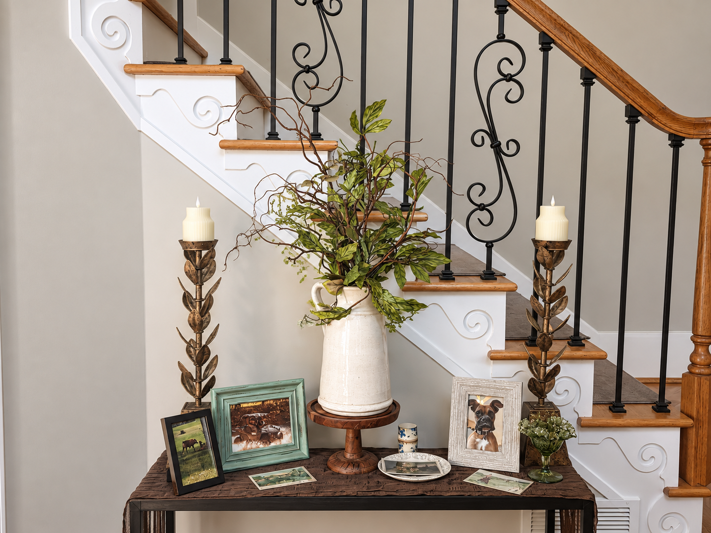 Entry console by staircase styled with greenery, framed wildlife art, candlesticks, and layered vintage pieces.