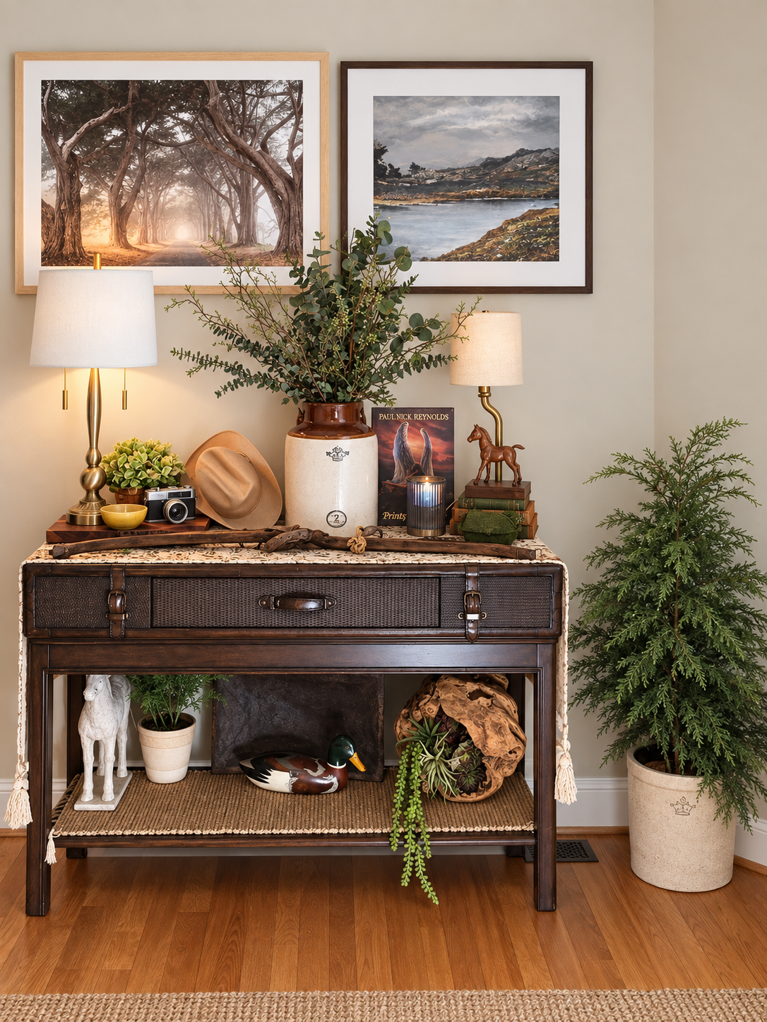 Dark wood console styled with framed landscape art, greenery, lamps, books, and collected décor.