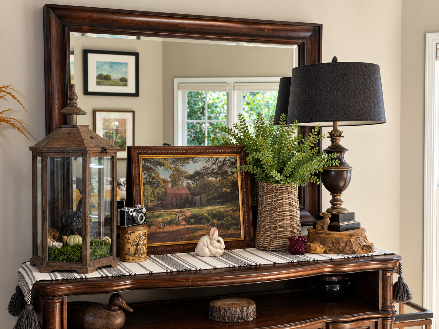 Decorative tabletop display with framed artwork, a lamp, a potted plant, a lantern, and small decorative items, reflected in a large mirror.