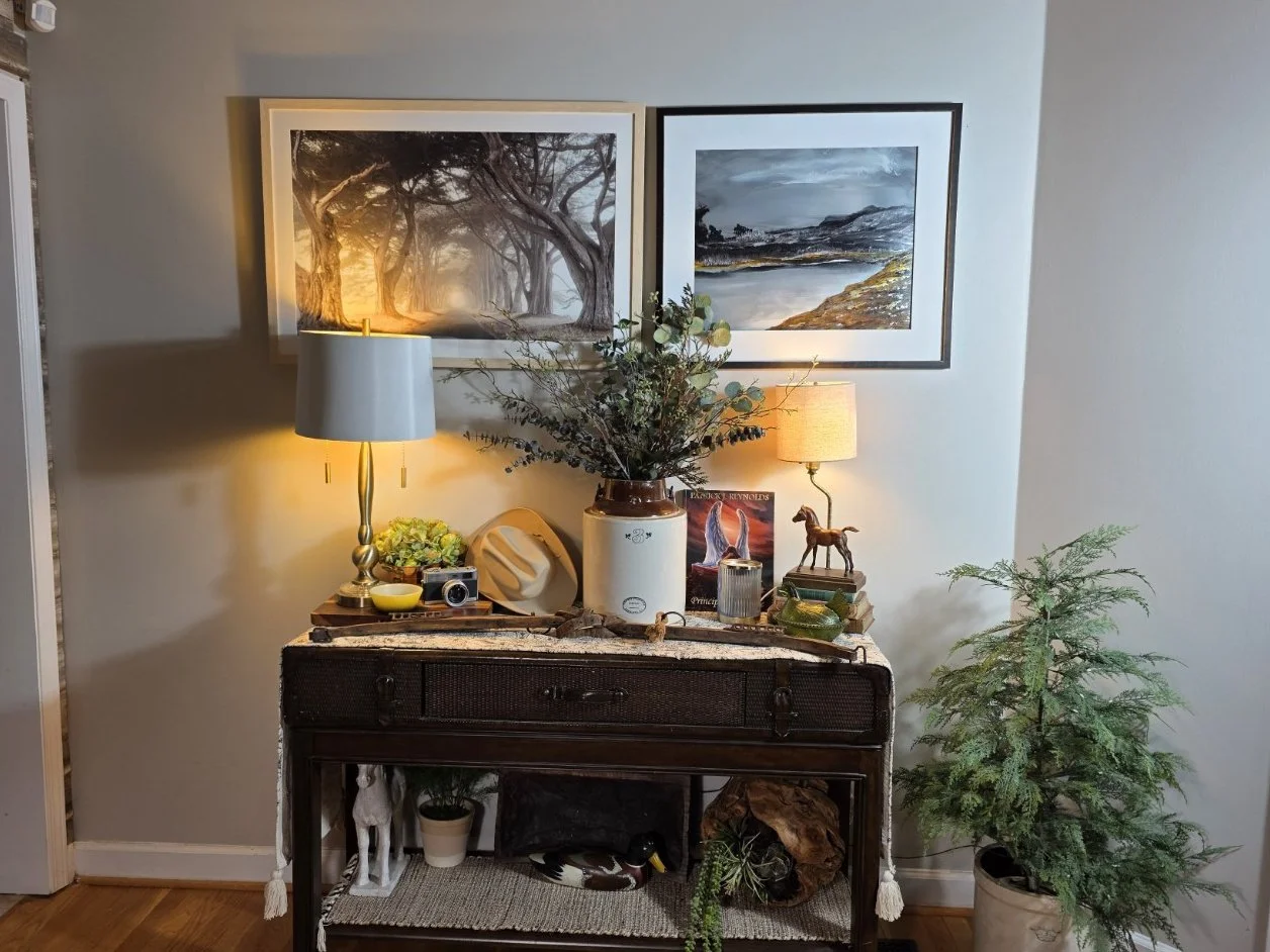 Decorative hallway table with two lamps, a large vase with greenery, and small decor items. Framed landscape photos on the wall, a small potted plant underneath, and a tree in a pot to the right.