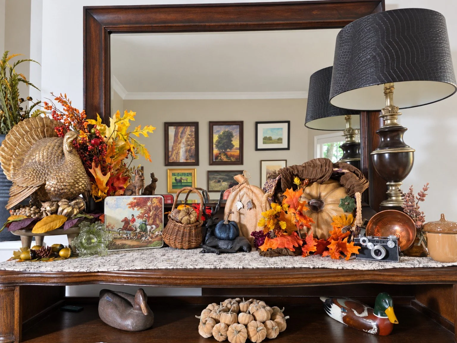Decorative fall display on a wooden table with pumpkins, leaves, animal figurines, and framed artwork in the background