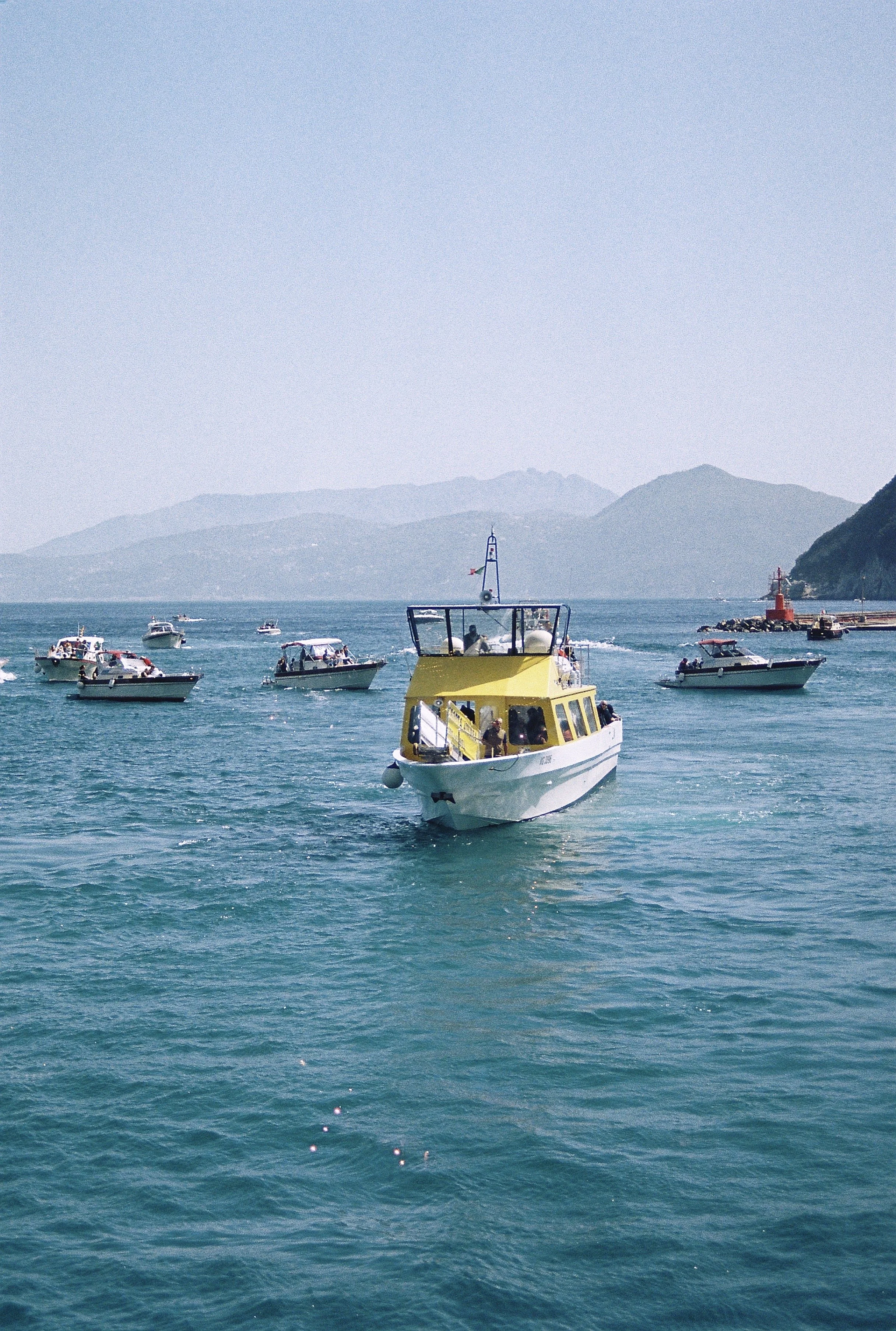 A yellow and white boat with a canopy sailing on turquoise water, surrounded by smaller boats, with distant mountains in the background.