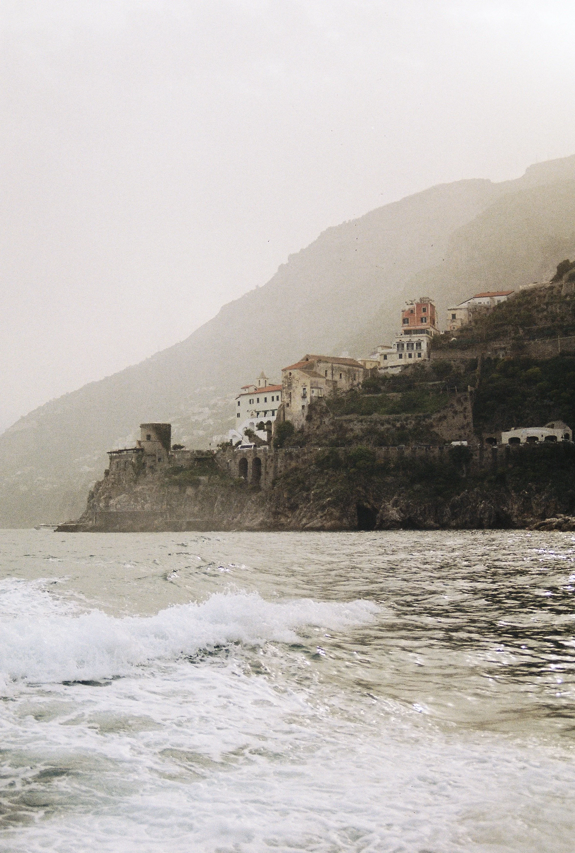 A coastal scene in Italy with waves in the foreground and a hillside with houses in the background, on an overcast day.