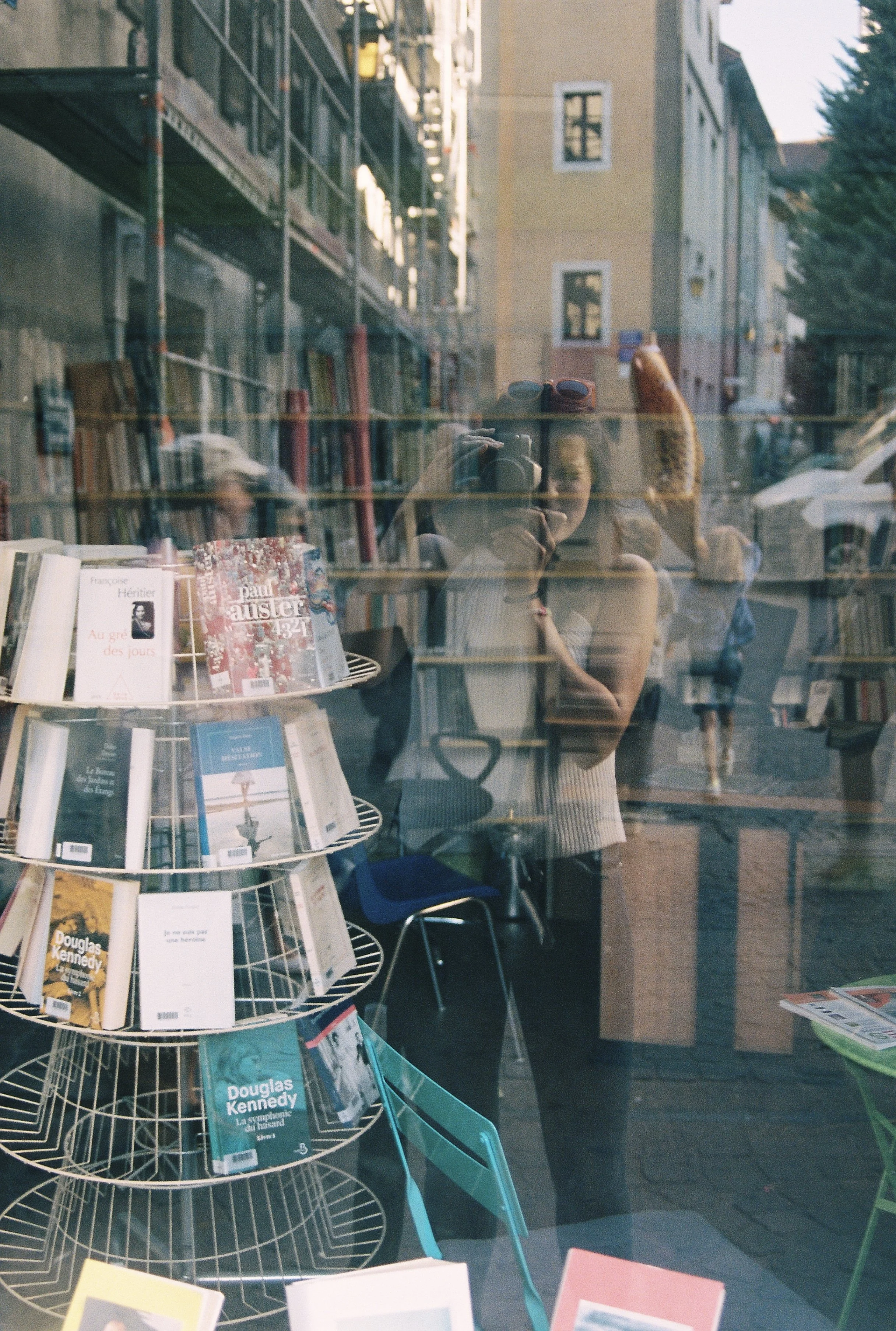 Books displayed on wire racks inside a bookstore, with reflections of buildings and people seen in the glass window.