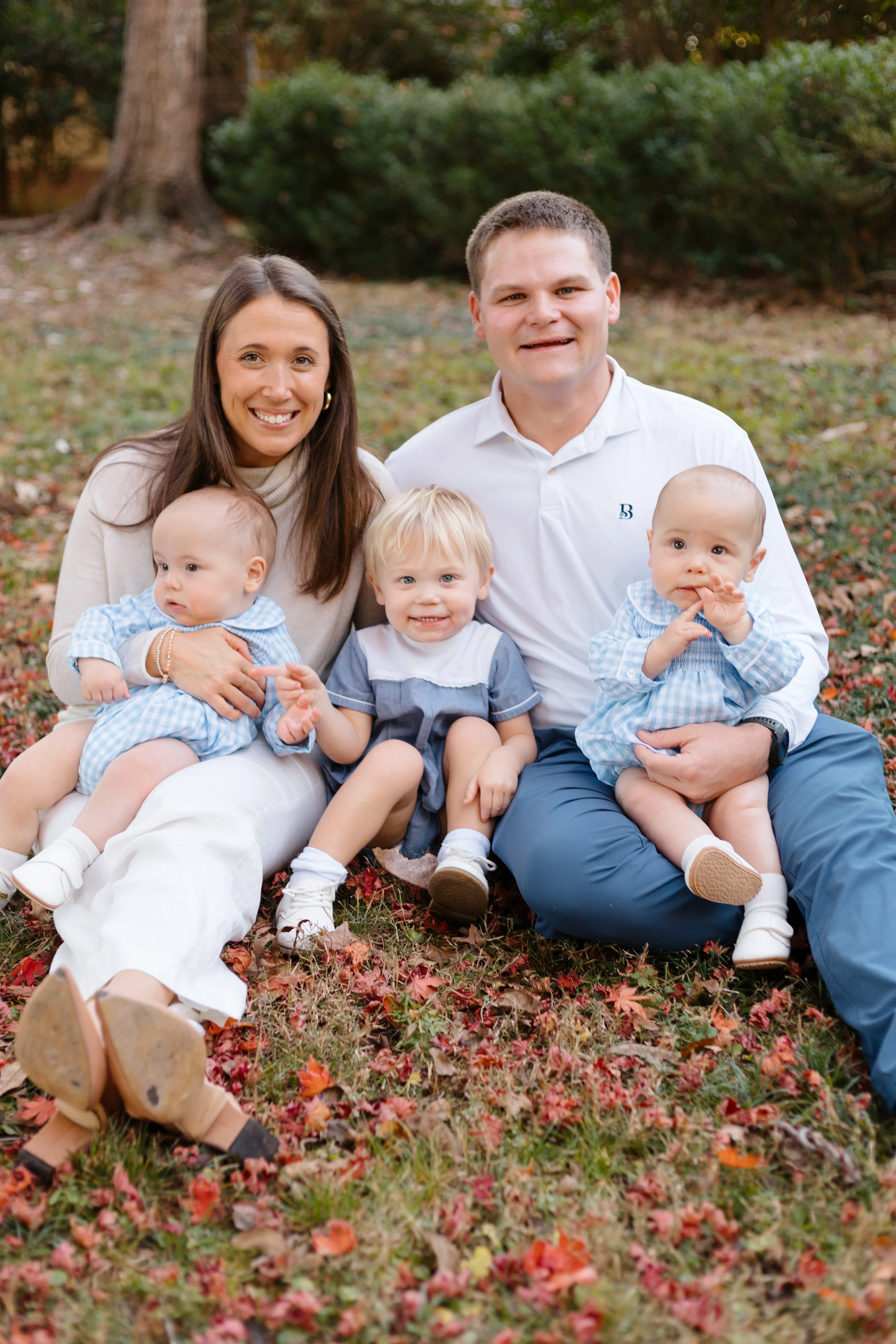 A family of five sitting outside on autumn leaves. Twin babies, a toddler, a woman, and a man, all smiling at the camera.