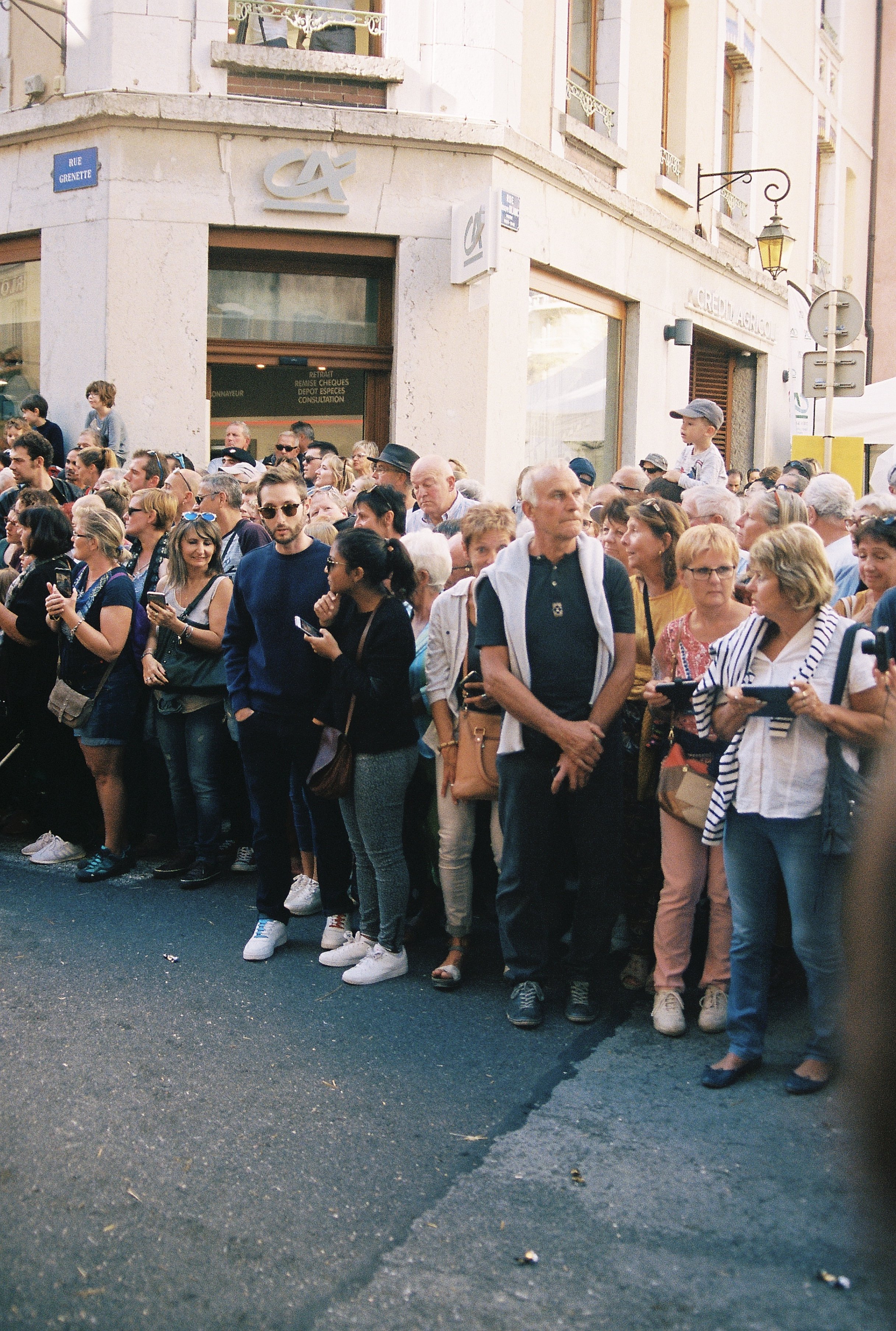 A crowd of people waiting in line on a city street. Some are on their phones, others are talking or looking around. The scene is in front of a building with a sign that reads 'Crédit Agricole' and another sign with the bank's logo. Taken in Annecy, F