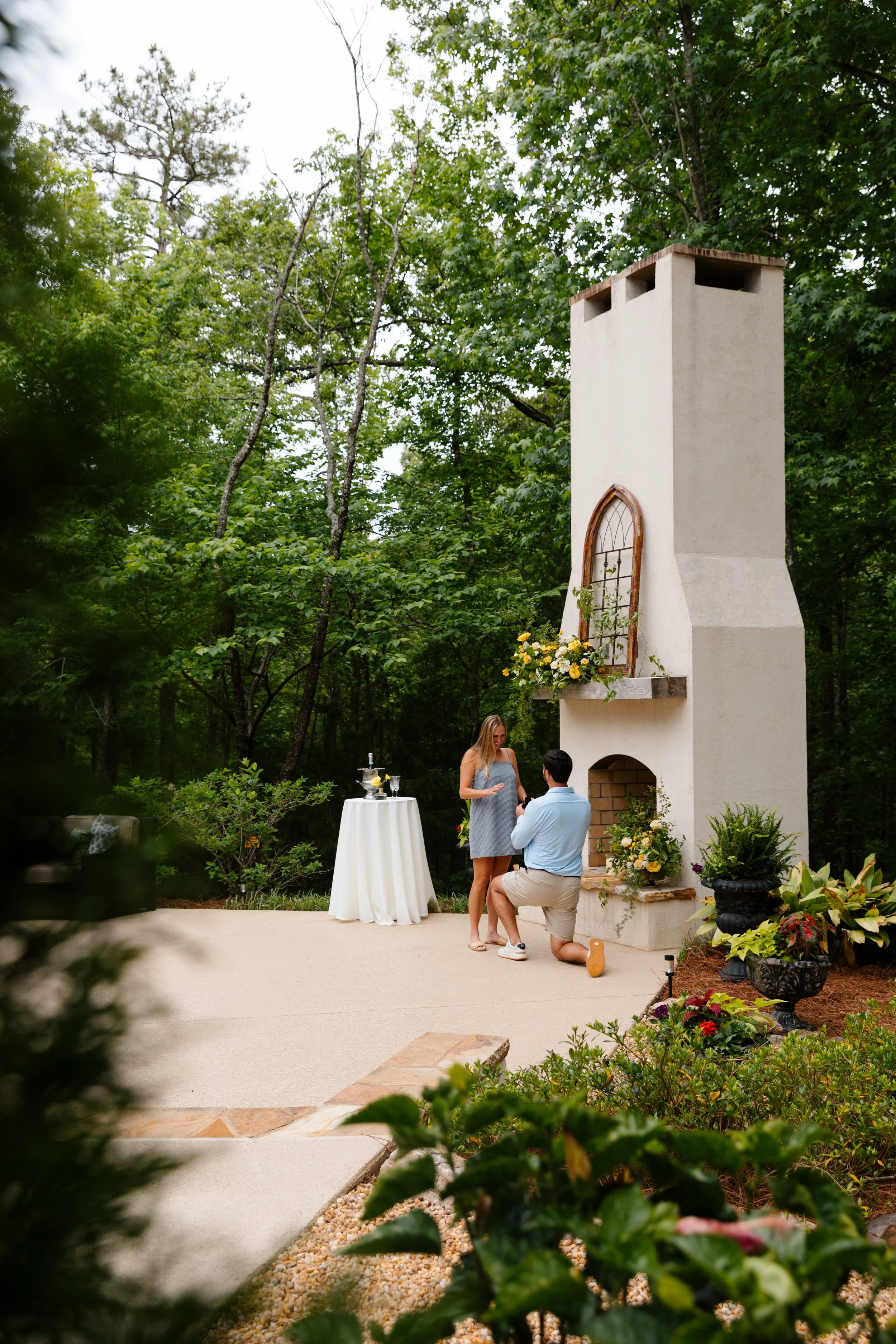 A couple is proposing marriage outdoors in front of a tall, white fireplace with a small table nearby. A woman is standing with her hand over her heart, and a man is kneeling on one knee. The scene is surrounded by greenery and decorative plants.