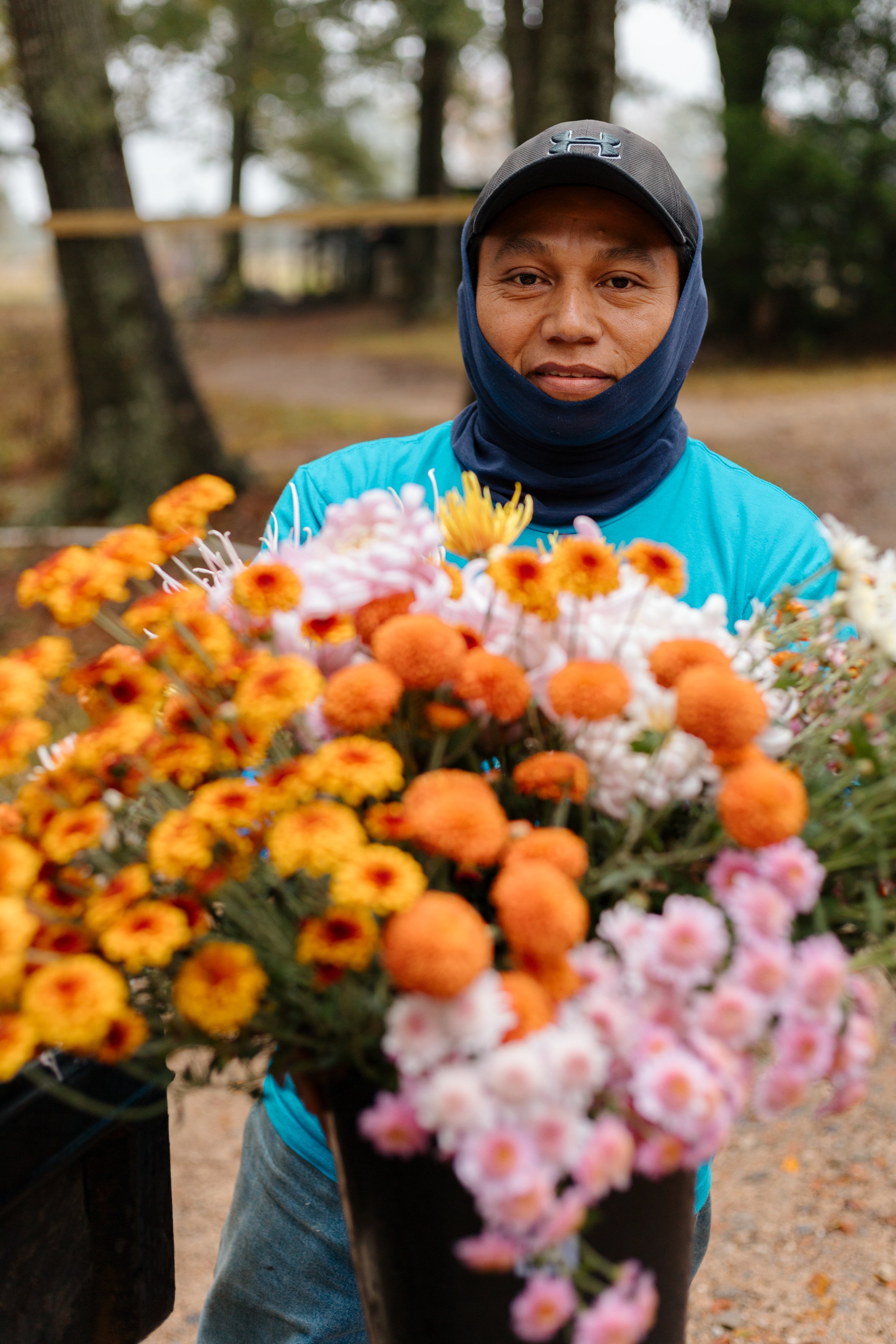 Local farmer outdoors holding a colorful bouquet of flowers, with a blurred background of trees and a fence.