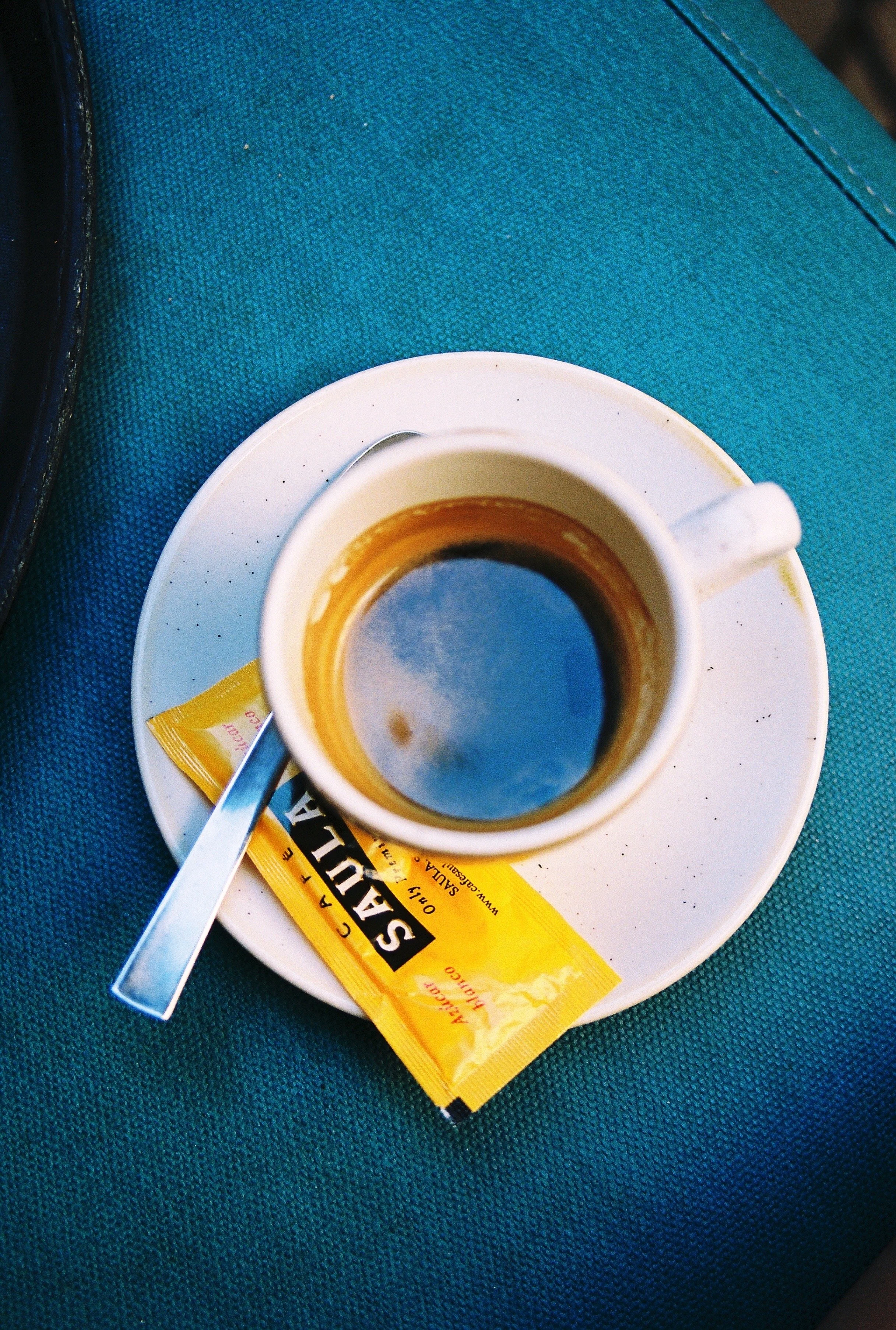 A small white cup of espresso on a saucer, with a silver spoon, on a teal surface. There is a yellow packet of instant coffee on the surface. Taken at a cafe in Barcelona, Spain.