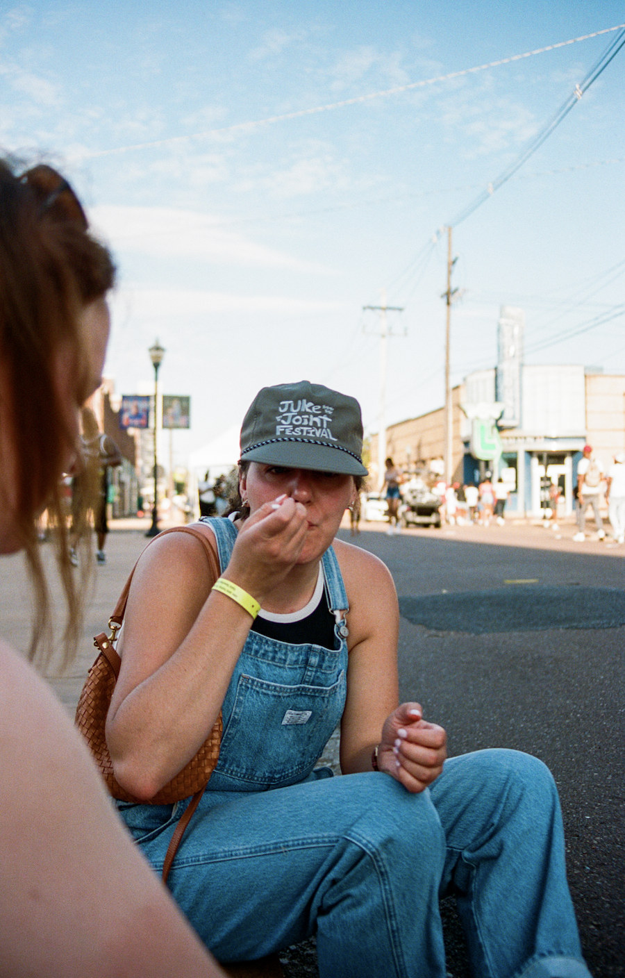 A woman in overalls and a "Juke Joint Festival" cap is sitting on the side of the street, eating tamales. The street is lively with people and storefronts in the background.