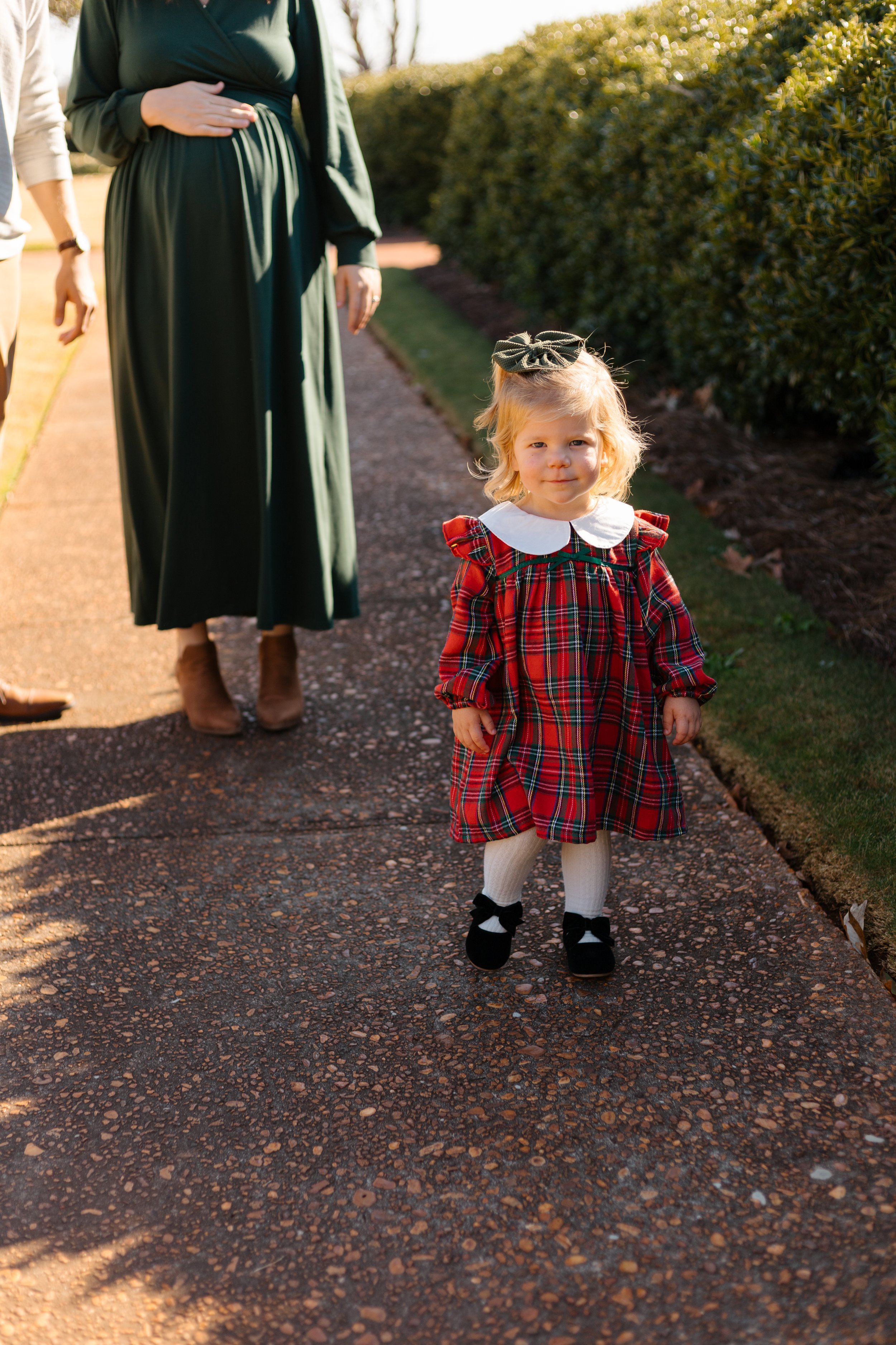 A young toddler with blonde hair, wearing a red plaid dress, white tights, and black shoes with bows, stands outdoors on a gravel path. She has a large bow in her hair and is looking directly at the camera with a slight smile. Behind her, adults are 