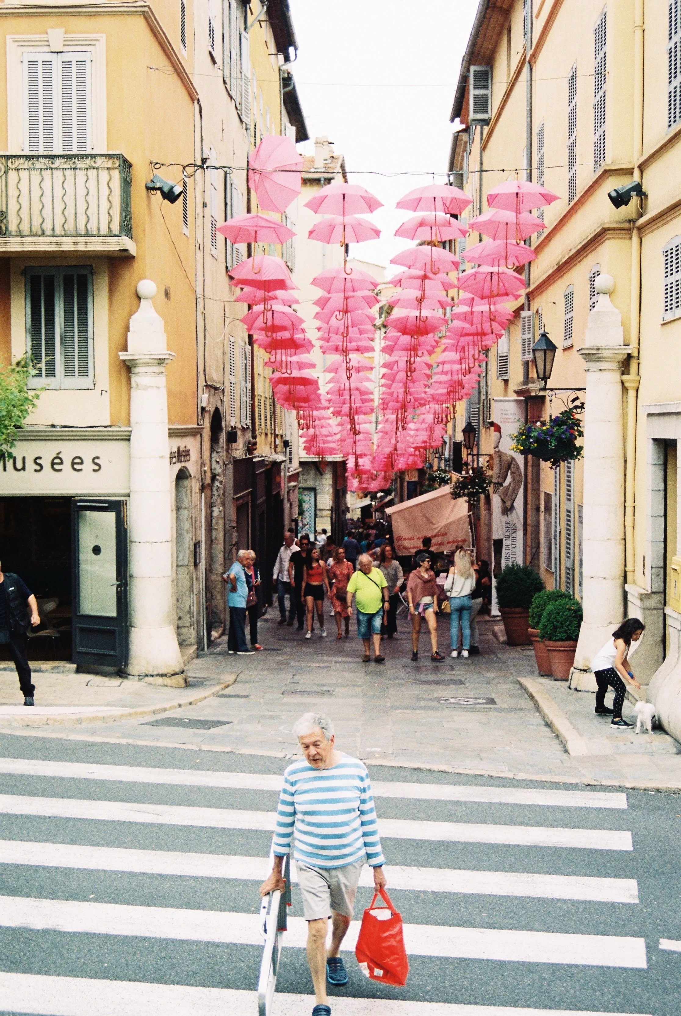 Street scene with several pink umbrellas hanging overhead, people walking on the street, some crossing a crosswalk, and buildings with yellow and beige facades on either side. In the South of France. 