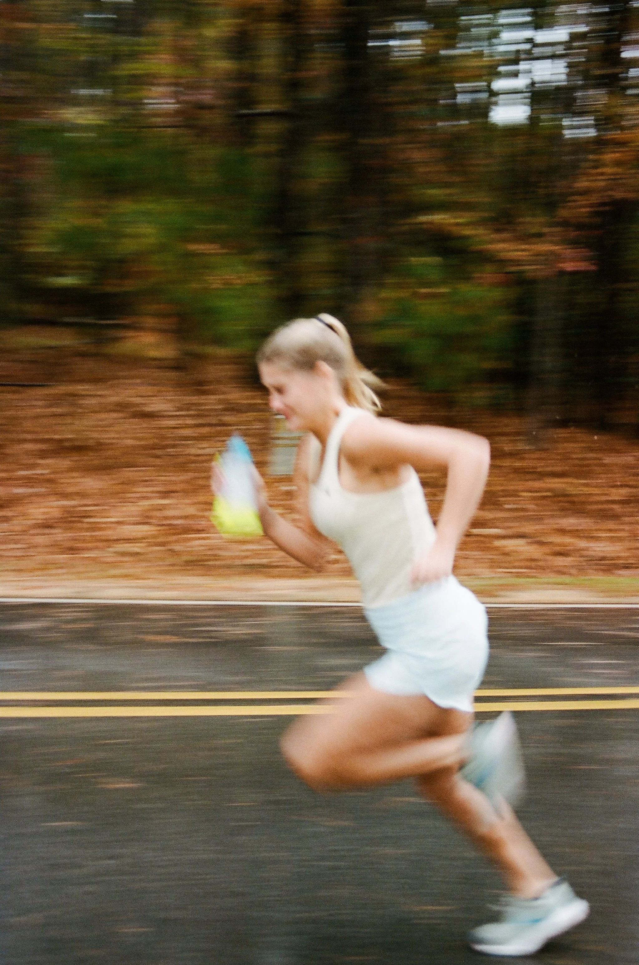 A woman running outdoors on a paved road, holding a water bottle, with a forest background and autumn leaves on the ground. Blurred motion indicates speed. Film photo for a small business. 