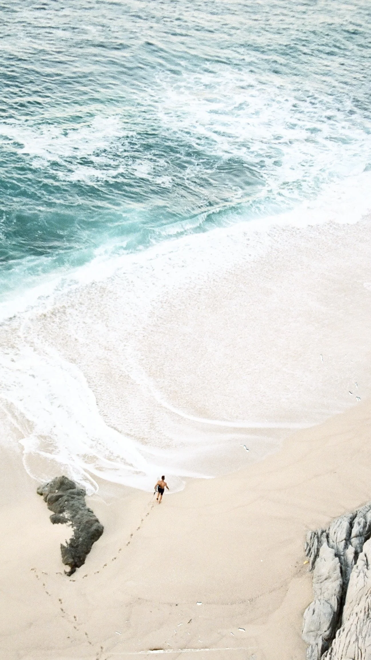 Aerial view of a person walking along sandy beach with large rocks, heading towards calm ocean waves. The sea is blue-green, and the sky is not visible.