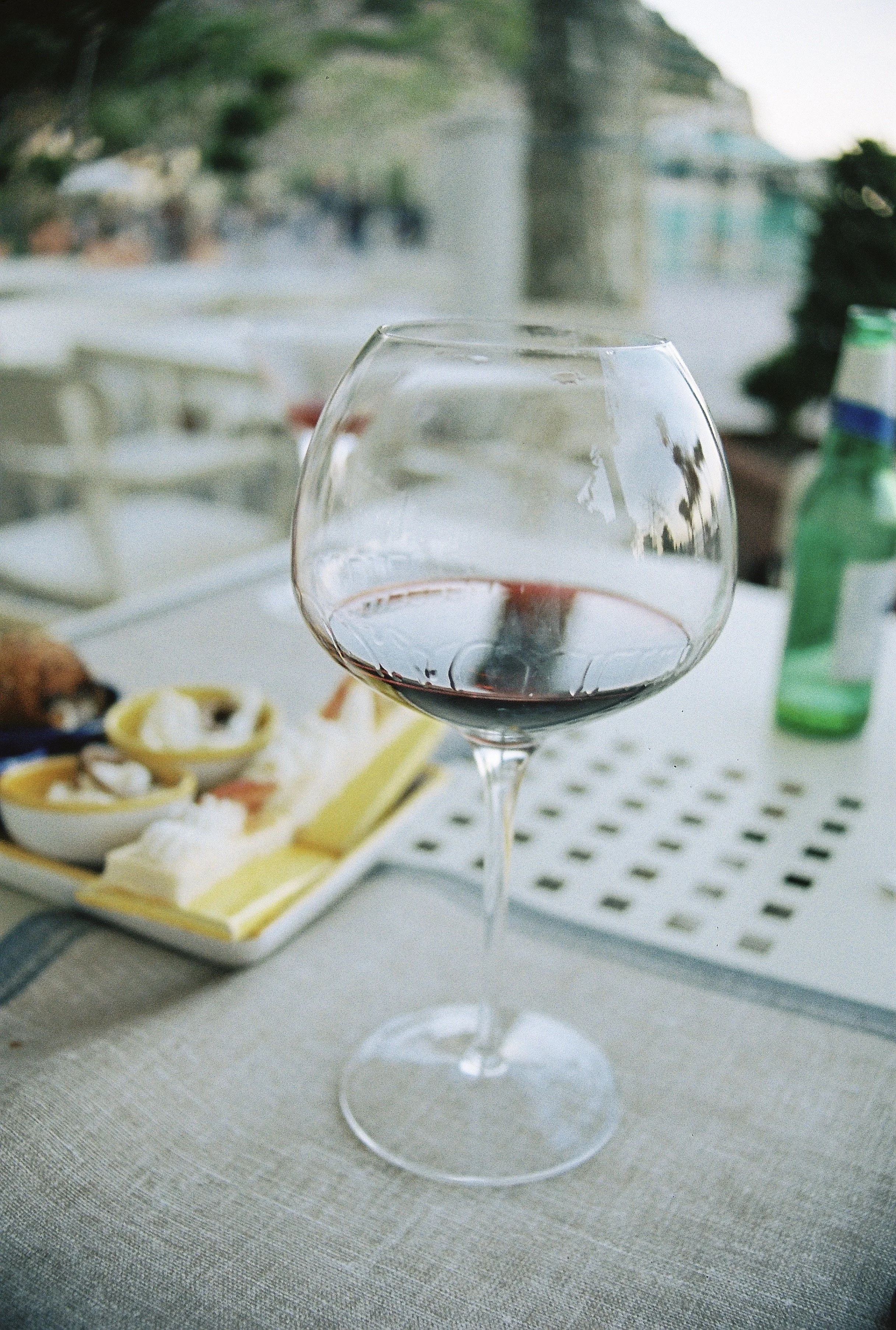 A glass of red wine on a table on the Amalfi Coast, with a blurred background showing a platter of cheese and lemon slices, a green bottle, and outdoors scenery.